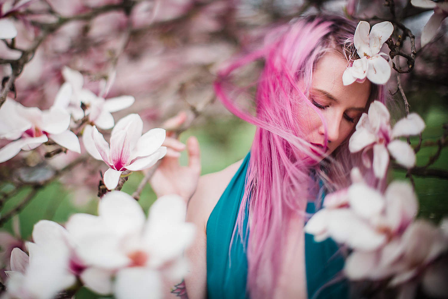 Photo of a young woman with pink hair and a teal dress standing in a tree of magnolia blossoms.