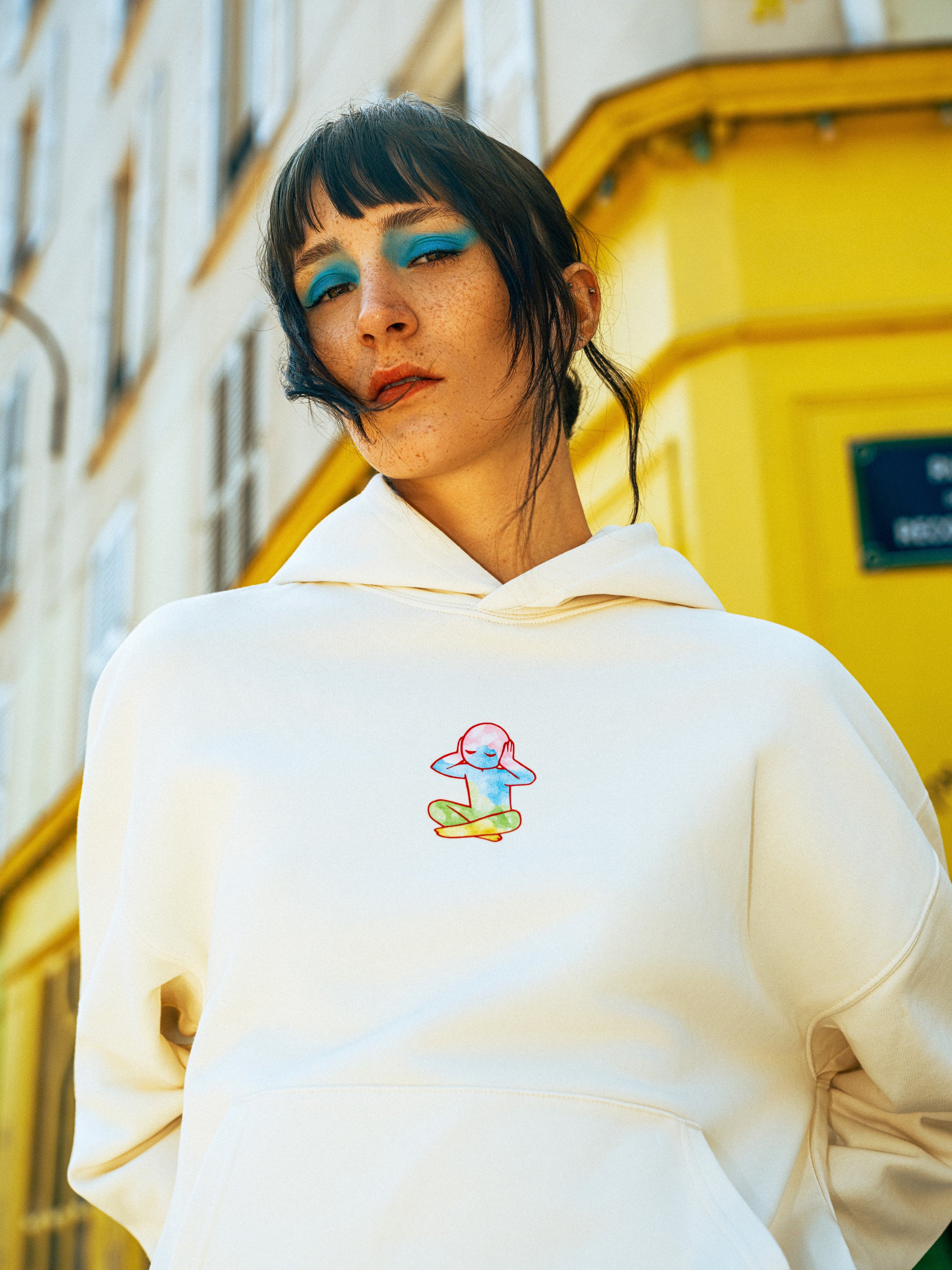 A woman with short black hair and blue eyeshadow wearing a white hoodie with a colorful meditating mascot on it, standing outdoors in front of a yellow building.
