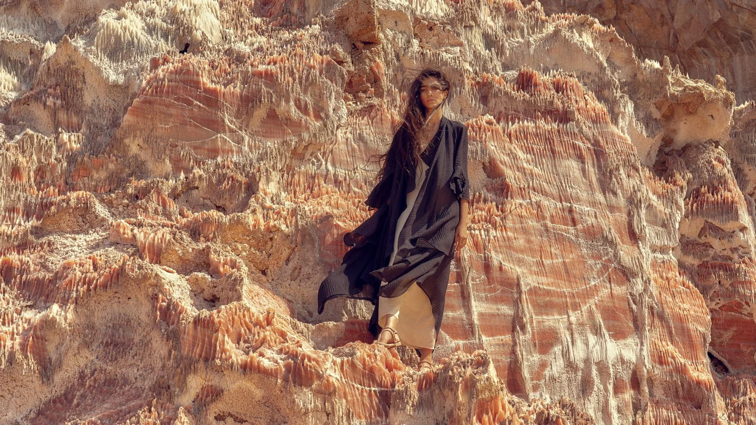 A woman in black and beige clothing standing on colorful, textured rock formations in a desert-like landscape.