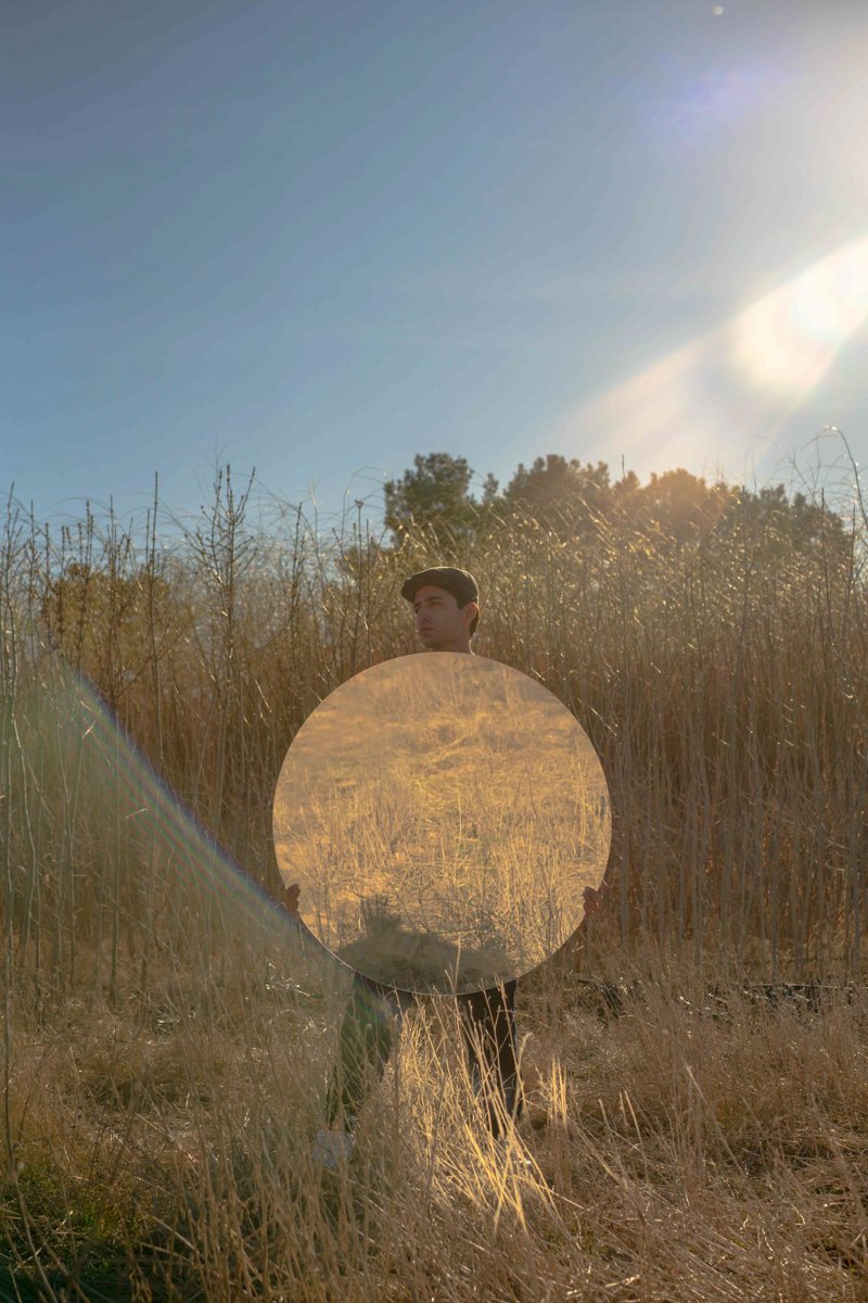 Person standing in a field holding a circular mirror reflecting dry grass, with the sun shining brightly and blue sky in the background.