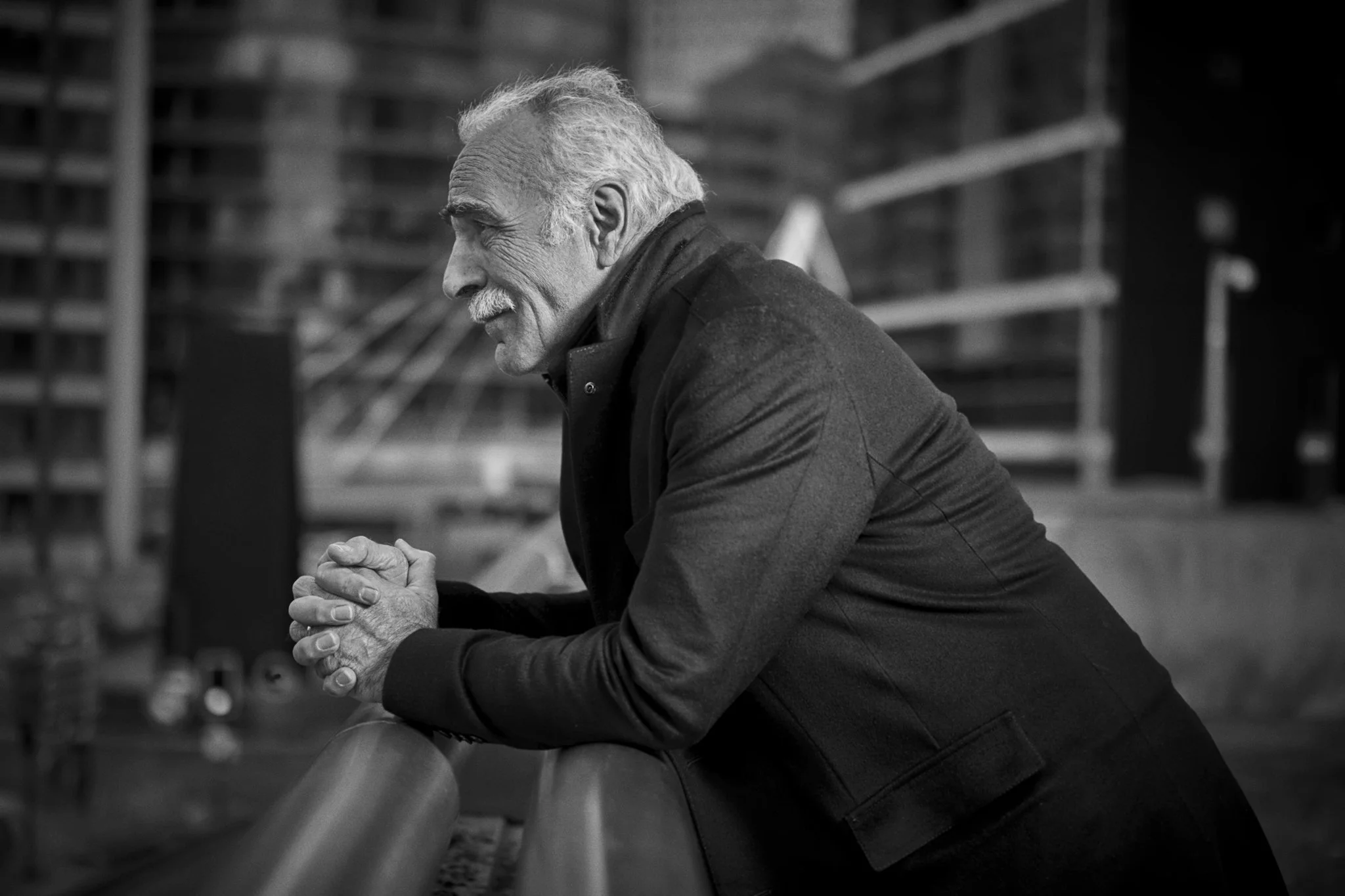 A black and white photo of an elderly man with gray hair and a mustache, sitting with his hands clasped in front of him, looking contemplative. The background shows a blurred industrial or warehouse setting with shelves.