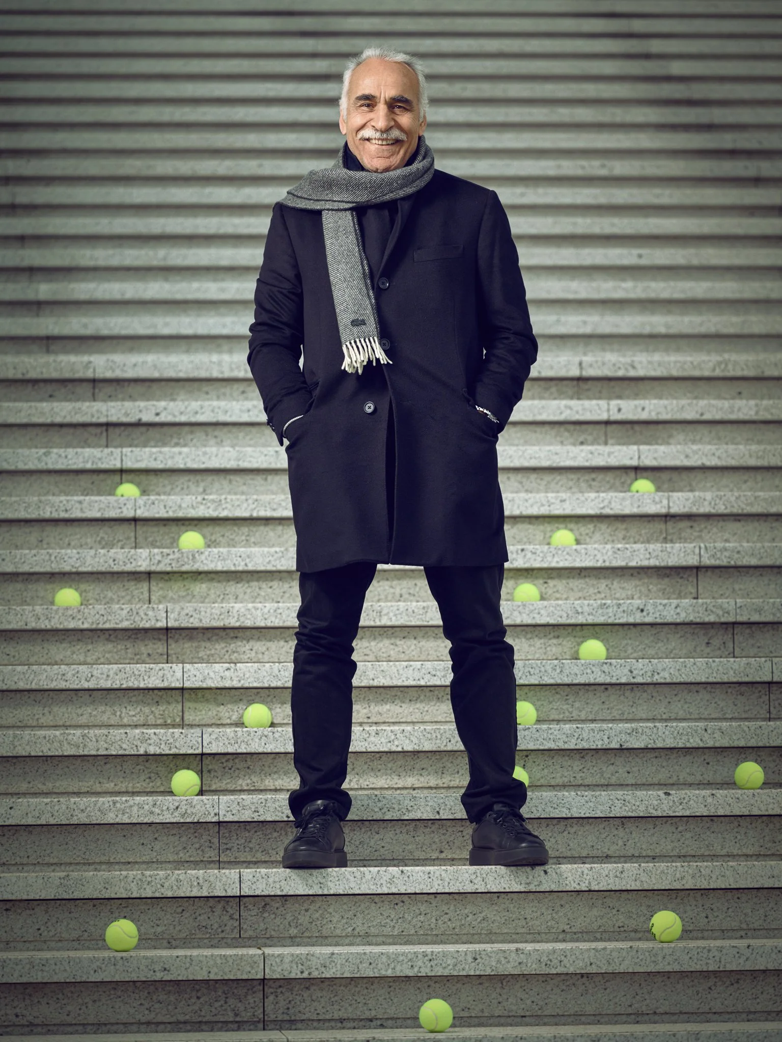 A smiling man with grey hair and a beard standing on concrete stairs, wearing a dark coat, black pants, black shoes, and a grey scarf, with tennis balls scattered around the stairs.