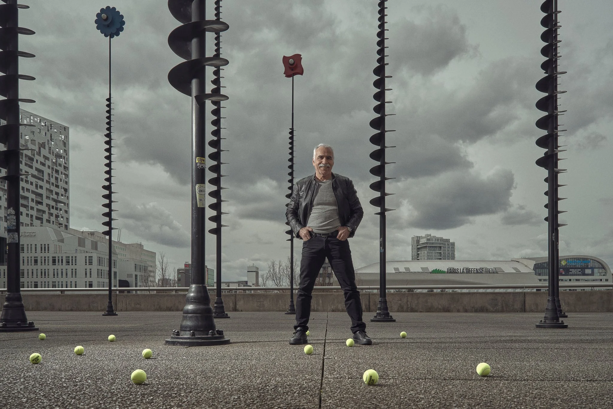 A man standing among tennis balls on a city street with tall lamp posts and modern buildings in the background, under a cloudy sky.