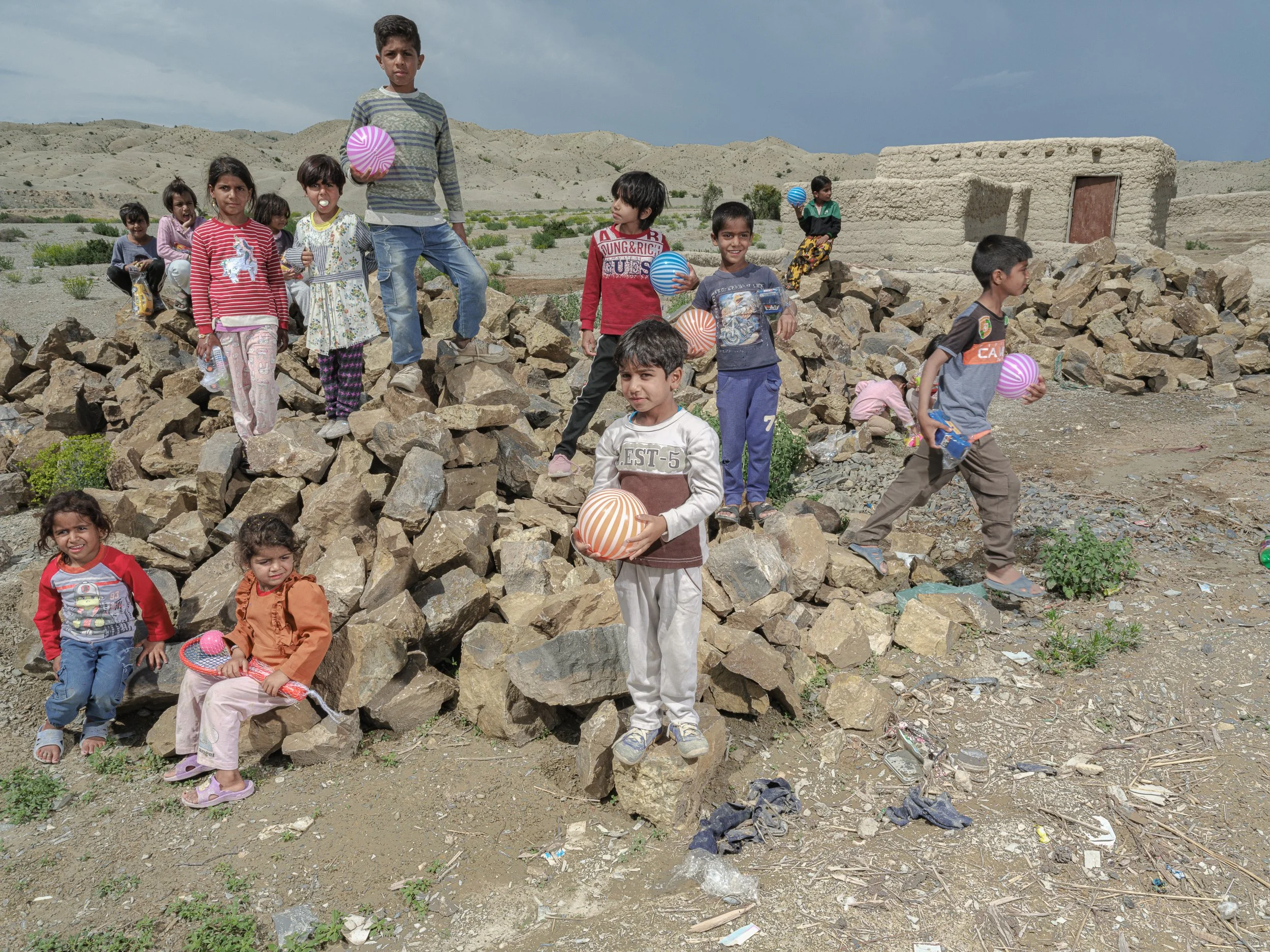 A group of children playing outdoors in a rocky, arid environment with a small adobe building in the background. They are holding balloons and wearing casual clothing.