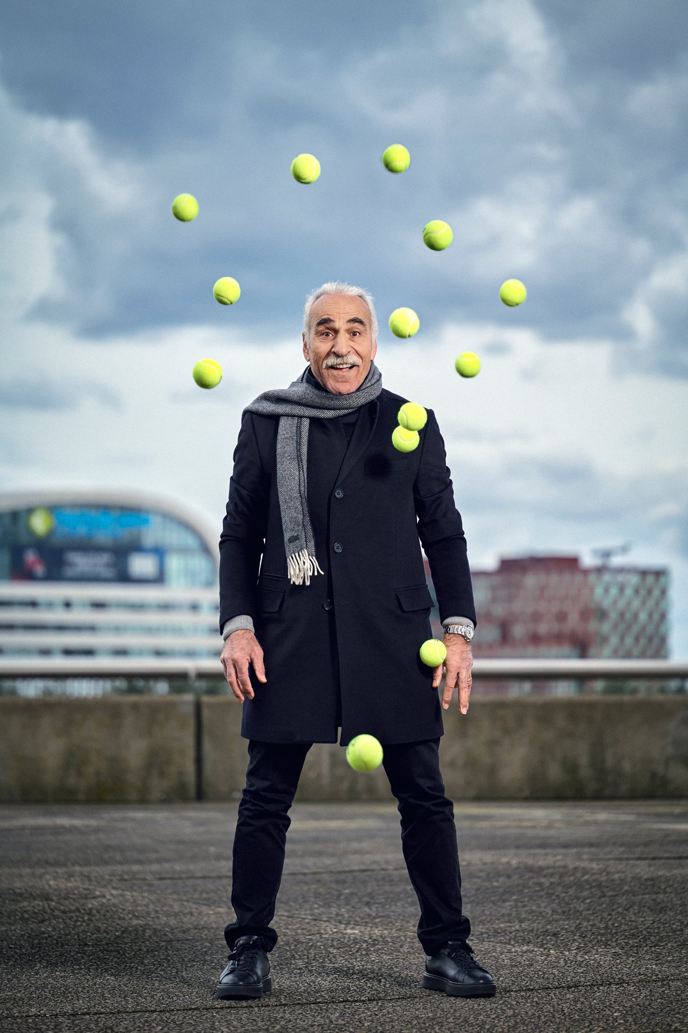 A man standing outdoors on a rooftop with a cityscape in the background, juggling tennis balls in the air, against a cloudy sky.
