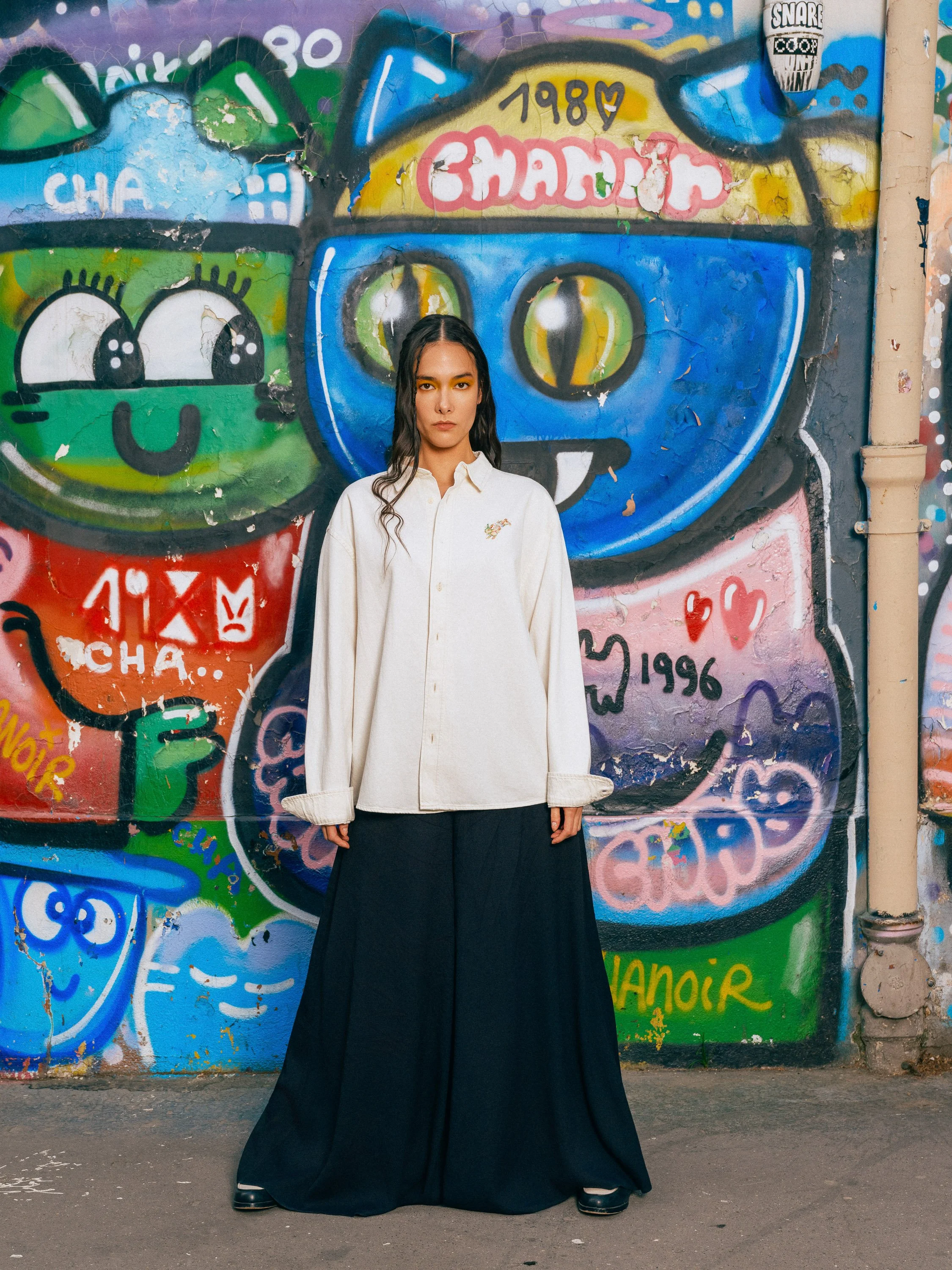 A woman with long dark hair wearing a white shirt and black skirt stands in front of a colorful graffiti wall.