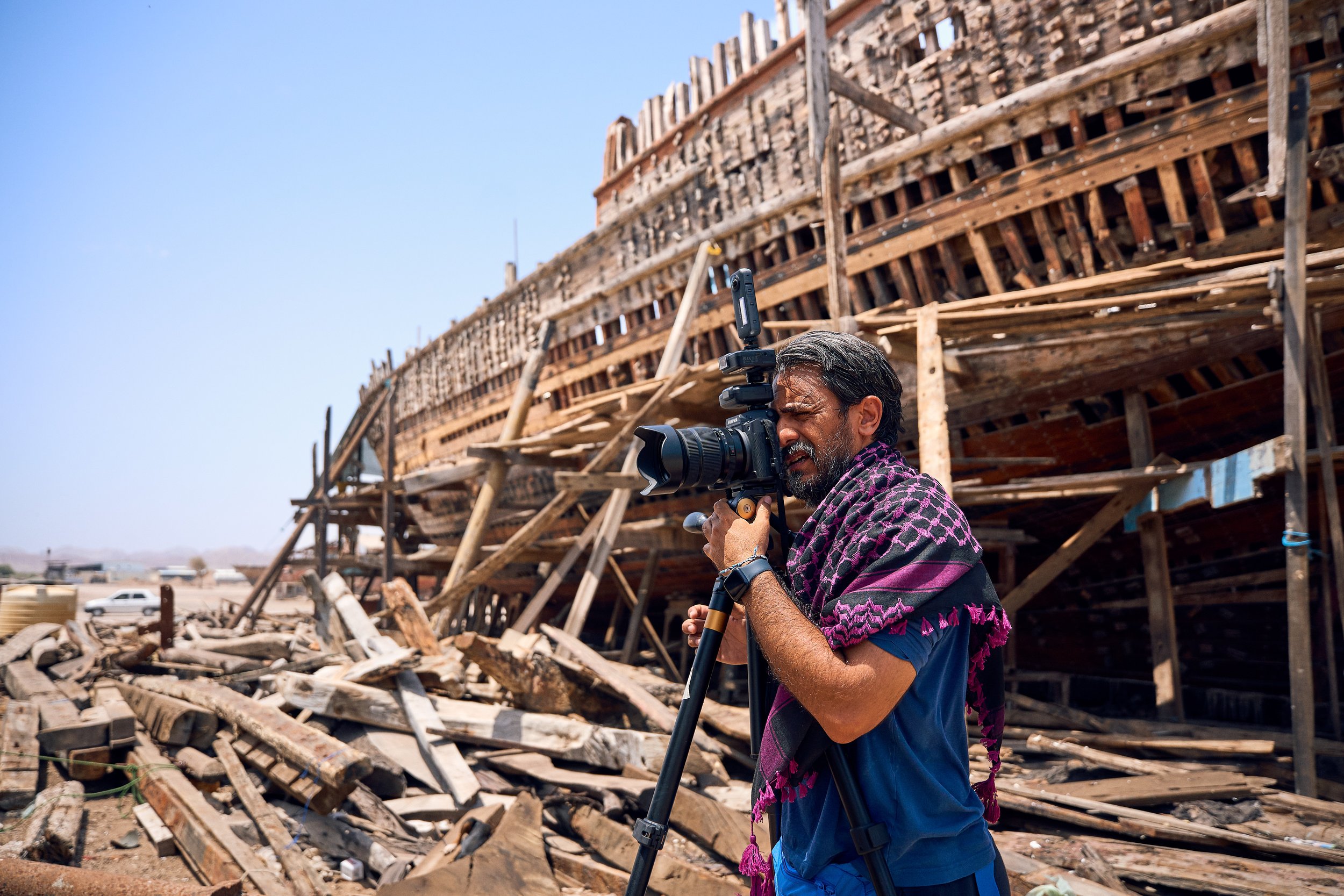 A man with a beard and dark hair, wearing a blue shirt and a purple patterned shawl around his neck, operates a professional camera on a tripod. Behind him is a damaged building made of wood and debris, with a clear blue sky overhead.