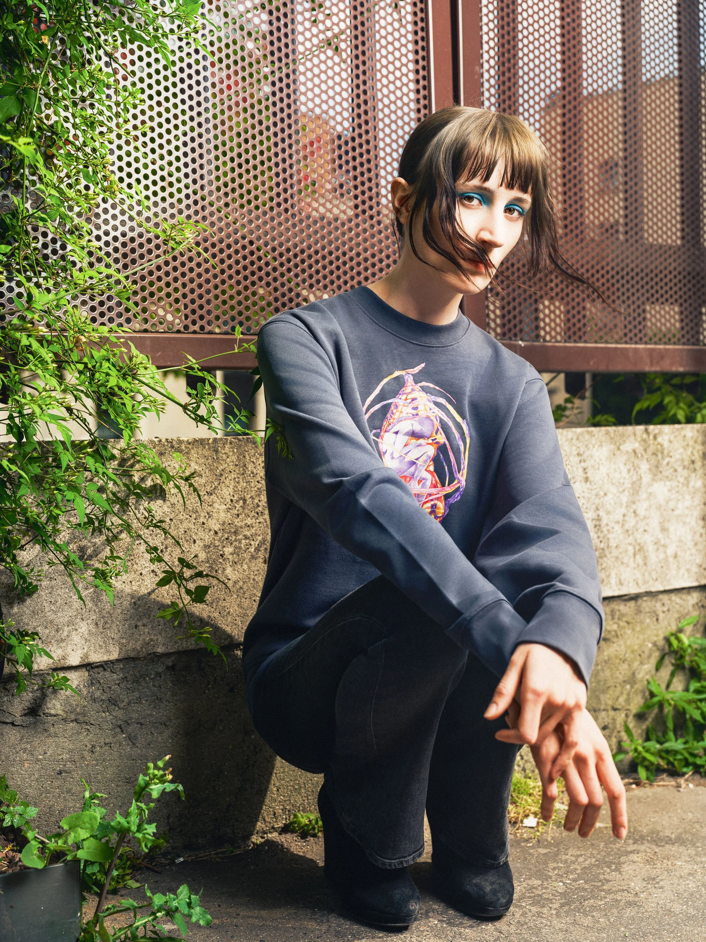 A young woman with short dark hair and blue eyeshadow crouching outdoors near a concrete wall with greenery, wearing a dark sweatshirt with a colorful skeleton design, and black boots.