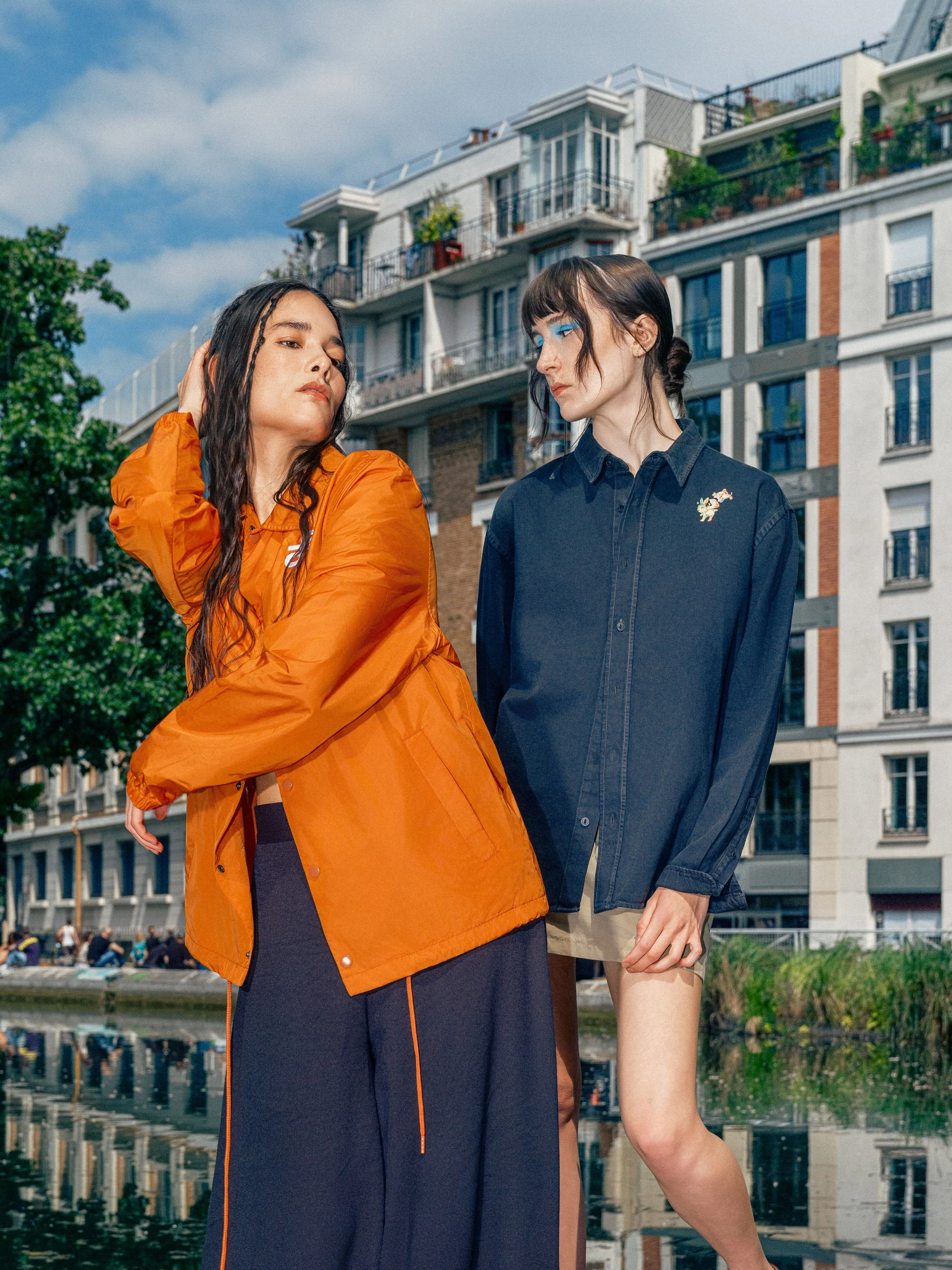 Two women standing outdoors near a body of water with city buildings in the background. One woman wears an orange jacket and black skirt, and the other wears a dark denim jacket and beige shorts.
