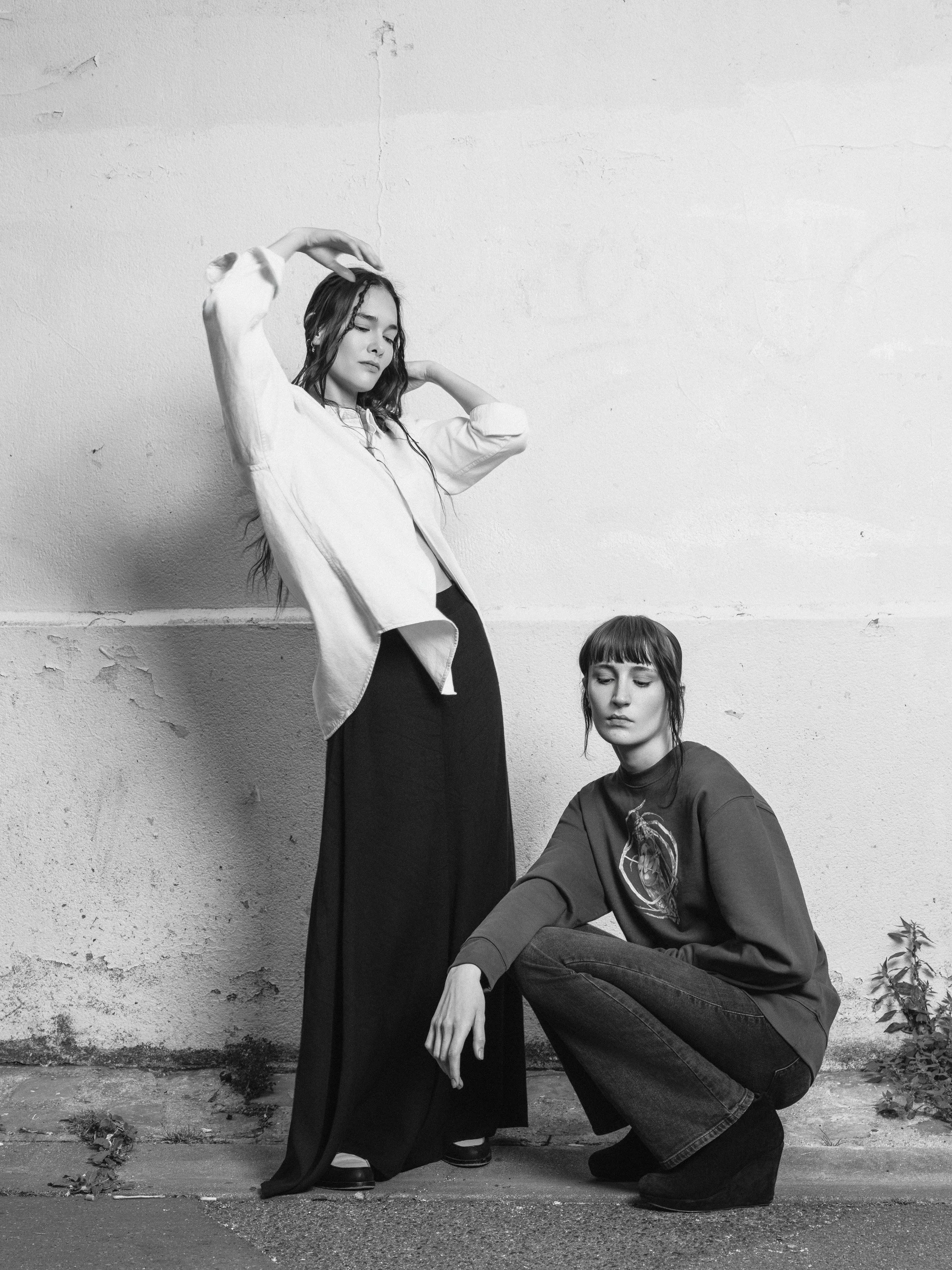 Black and white photo of two young women against a weathered wall. One woman is standing with eyes closed, wearing a loose blazer and long skirt, with one arm raised and hand resting on her head. The other woman is sitting on the ground with her head
