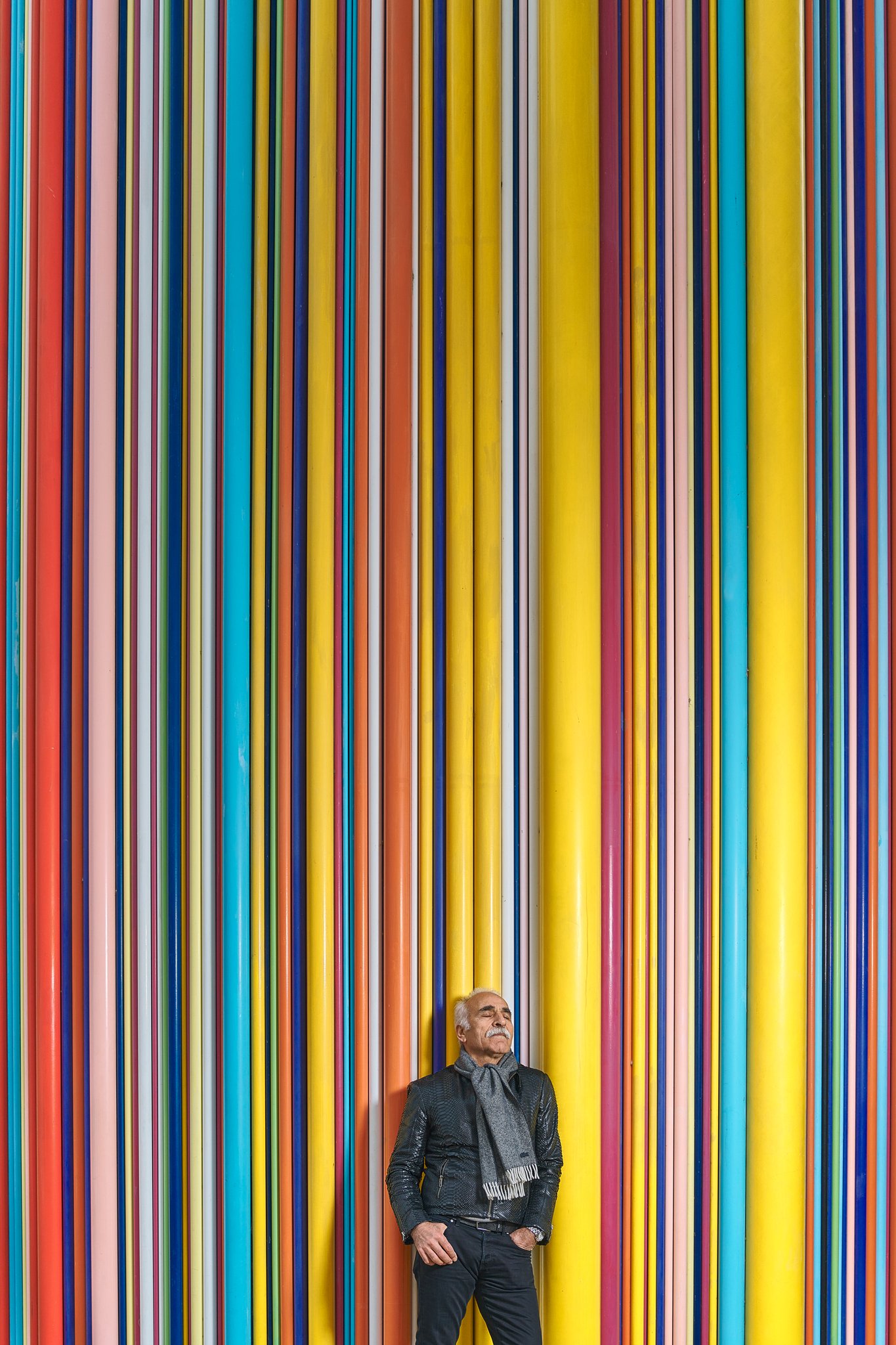 An elderly man with white hair and a mustache, wearing a black leather jacket and a gray scarf, is standing with eyes closed in front of a colorful vertical striped background at an art installation or event.