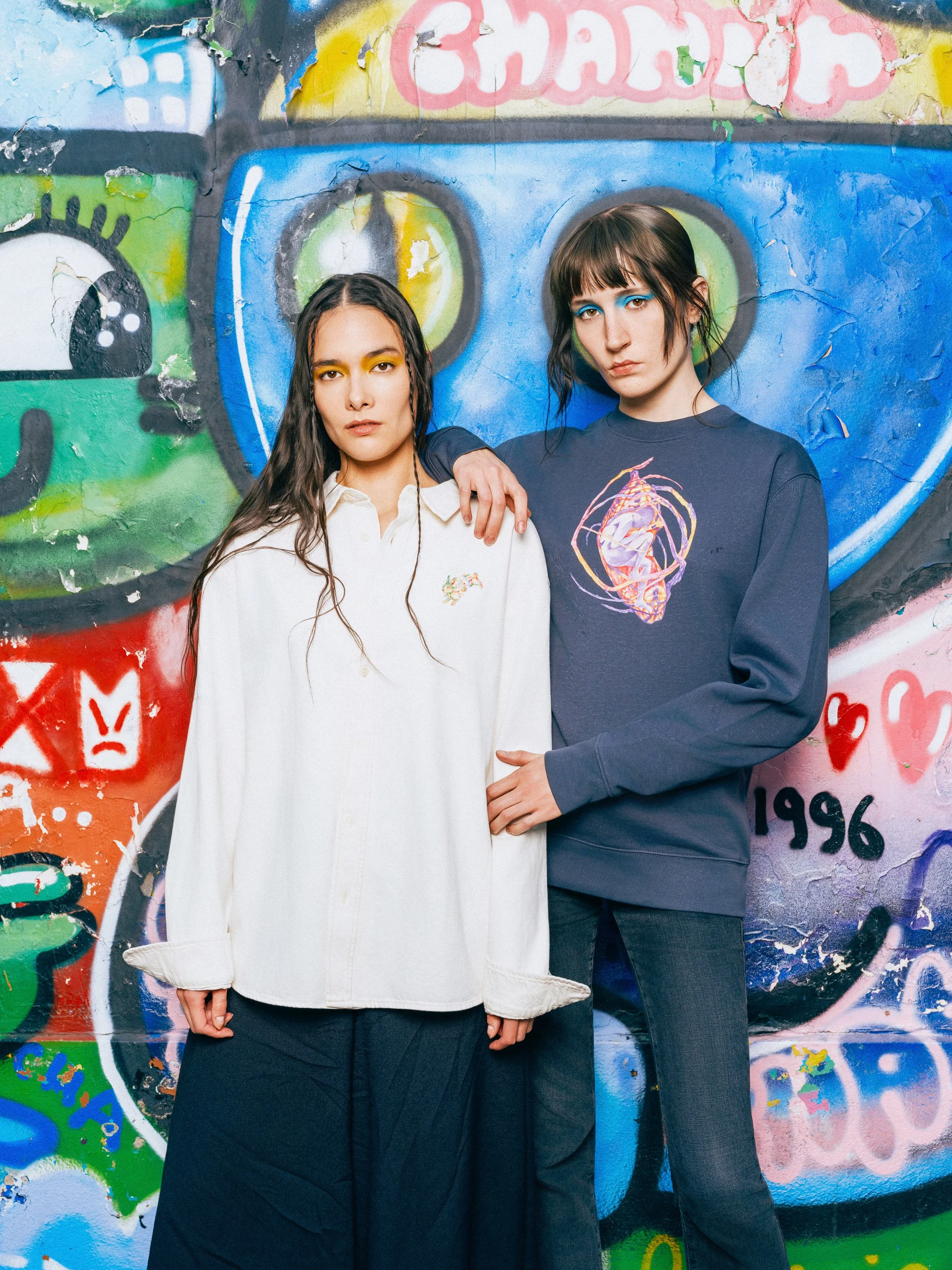 Two women with dark and light brown hair standing in front of a graffiti wall featuring vibrant colors, abstract shapes, and text. One woman has long, straight hair with yellow eye makeup, wearing a white shirt and black pants. The other woman has a 