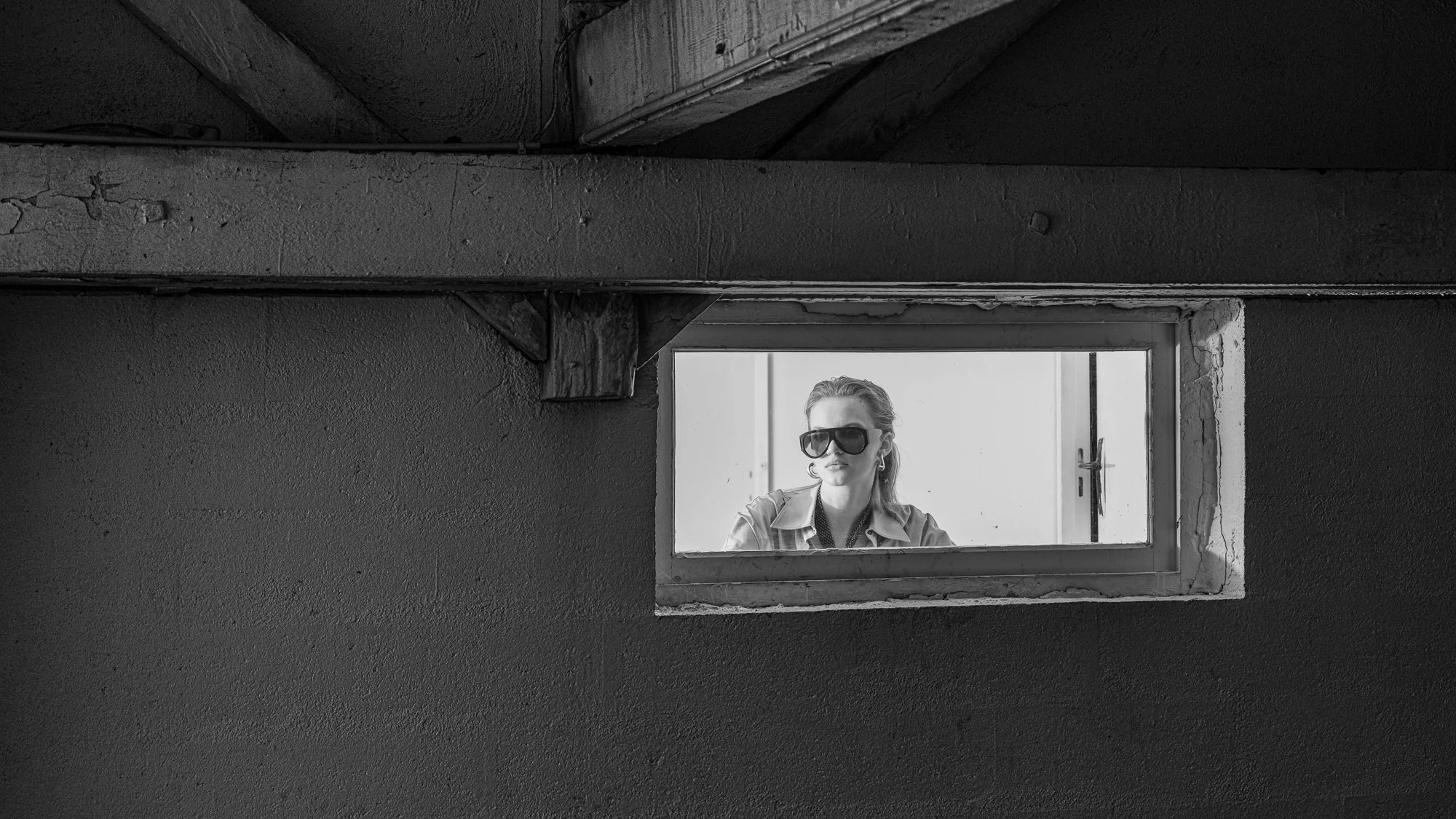 Woman wearing large sunglasses and earrings looking out of a small rectangular window in an unfinished, concrete wall.