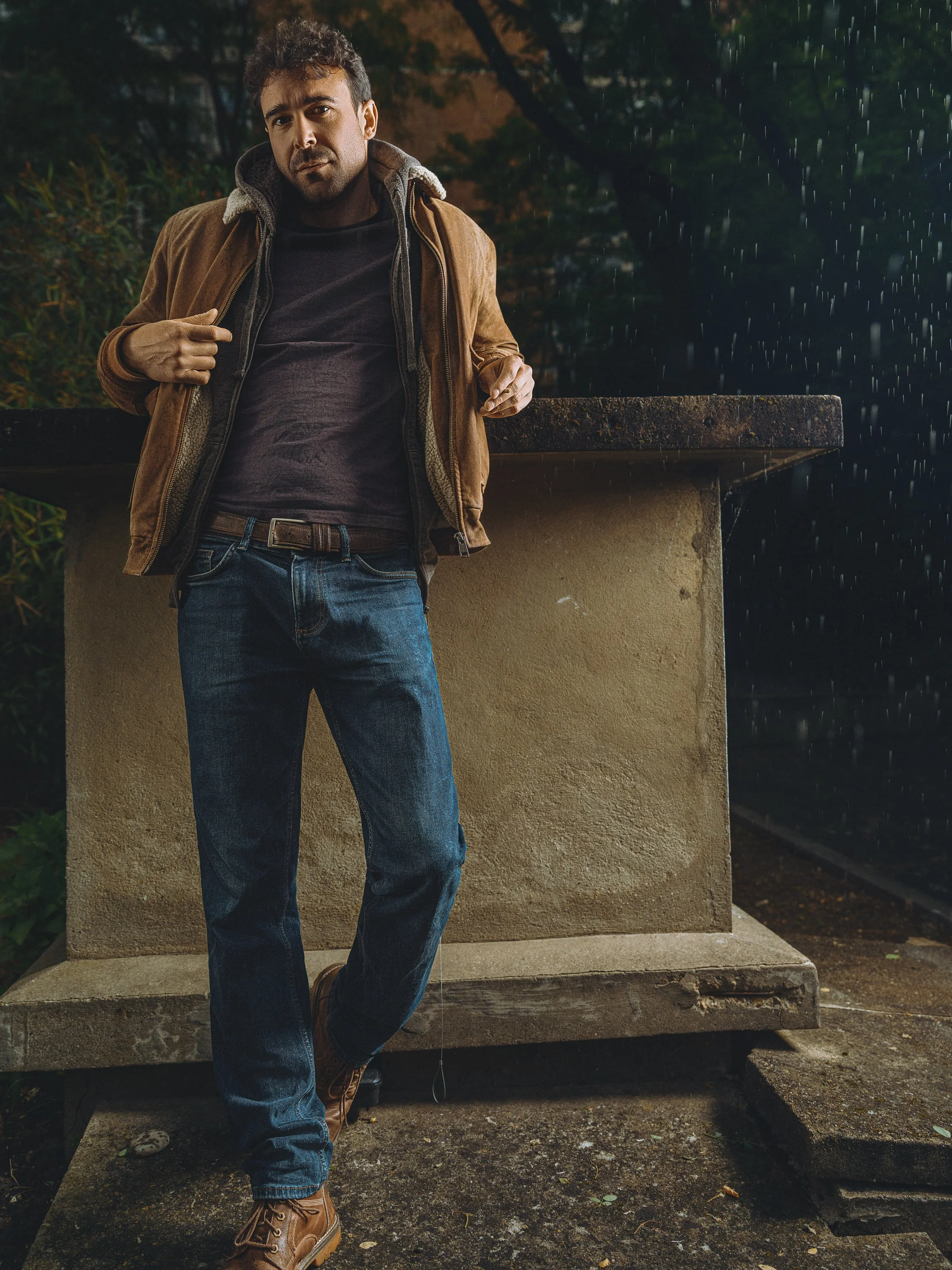 A man with dark, curly hair and a beard leaning against a stone ledge outdoors at night in the rain, wearing a brown jacket, dark shirt, blue jeans, and brown boots.