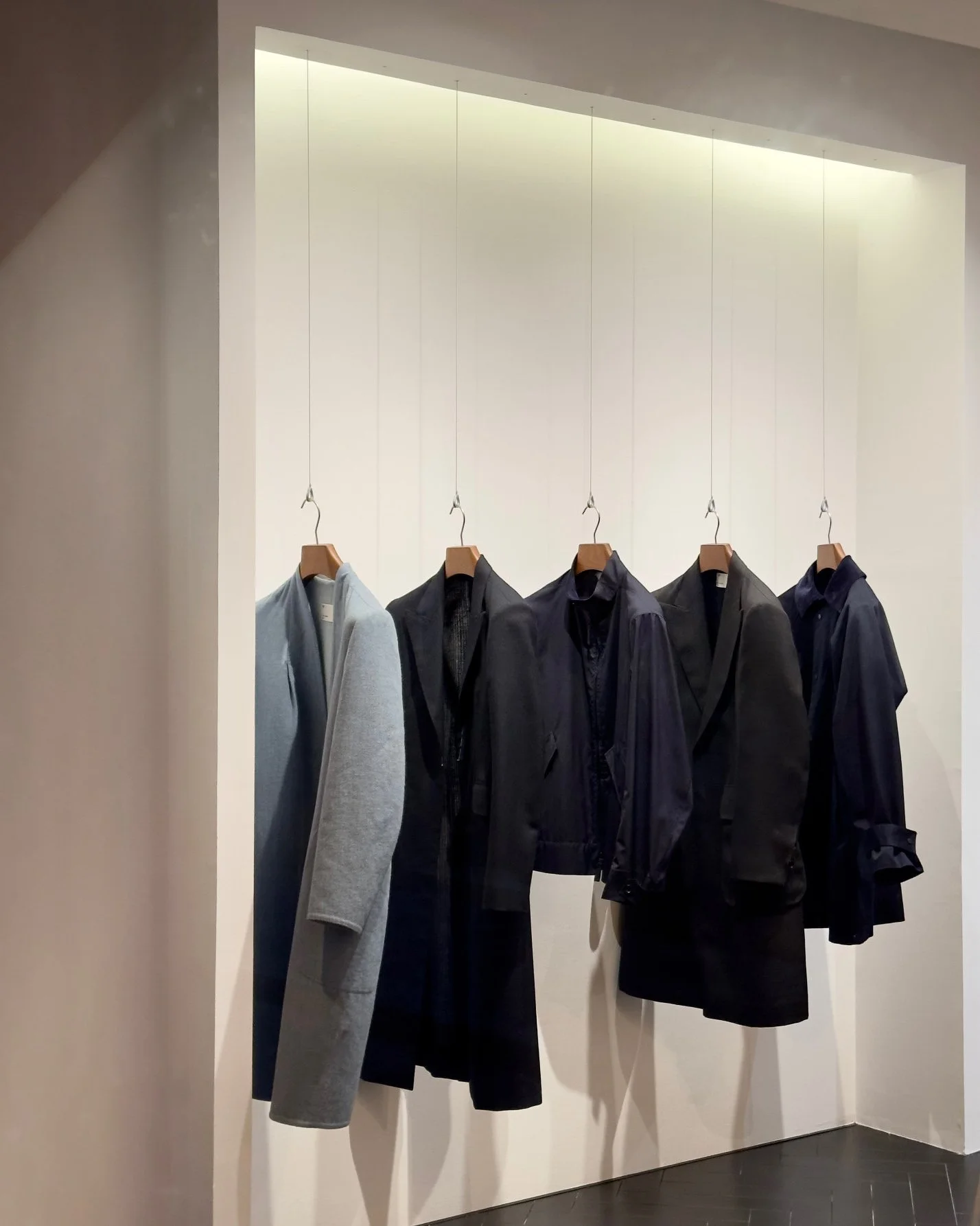 Vêtements pour hommes suspendus sur des cintres dans un magasin, incluant un manteau gris, plusieurs vestes noires et des chemises, présentés dans une zone bien éclairée