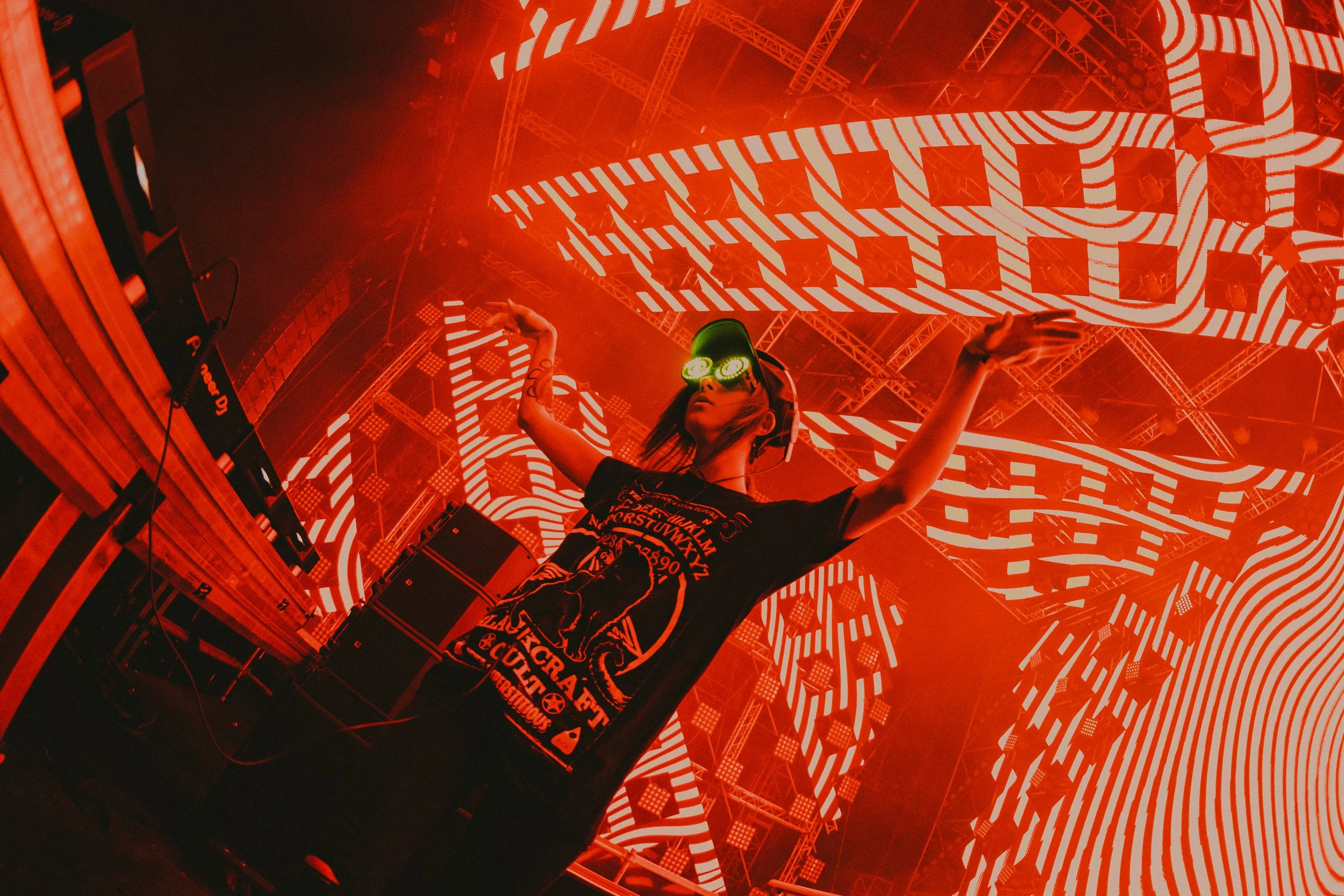 A DJ with glowing green glasses and a black t-shirt performing on stage with red and white LED lights and abstract patterns in background.