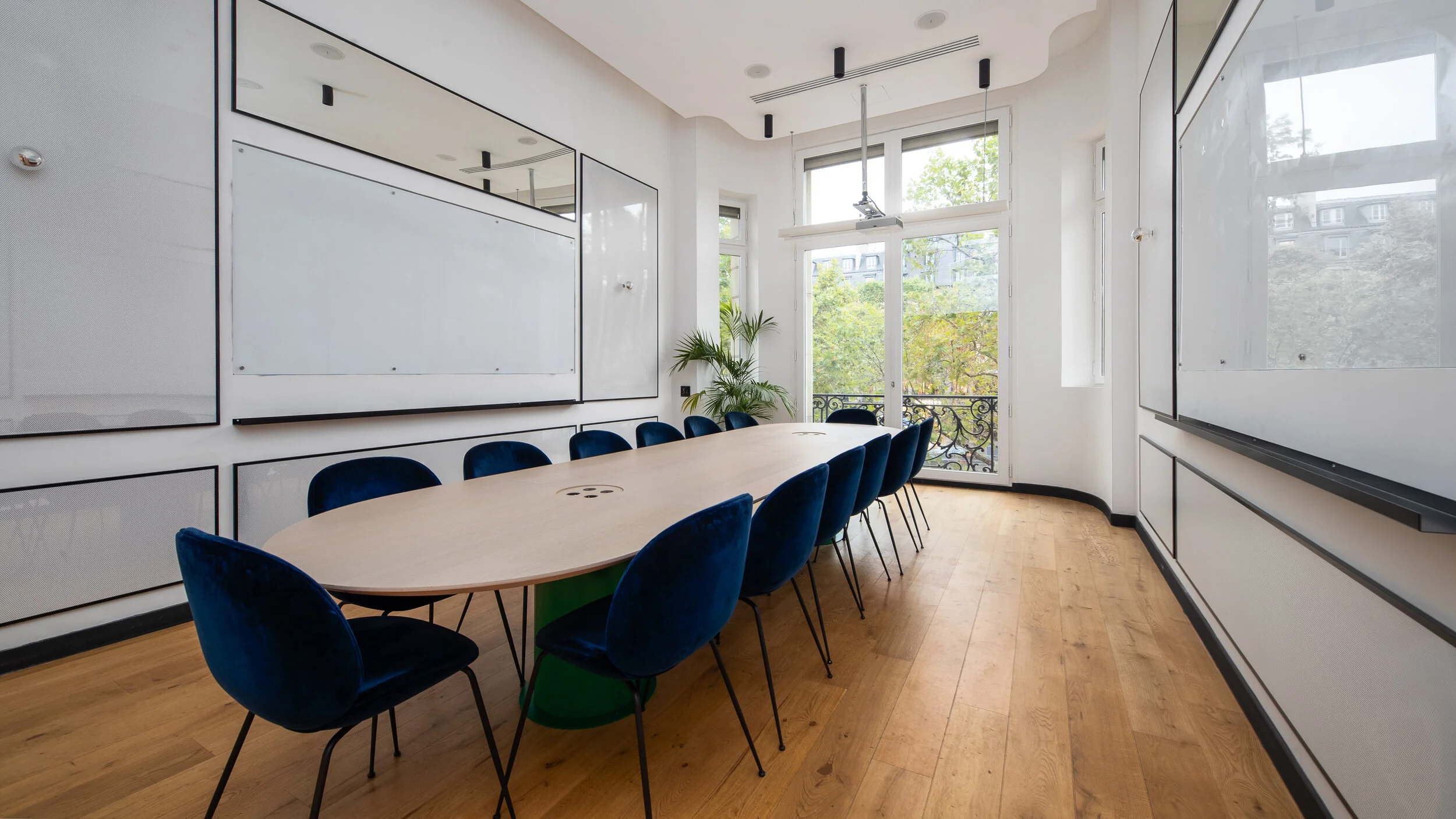 Modern conference room with a long oval table surrounded by blue velvet chairs, large windows with green trees outside, white walls with black trim, and a whiteboard on the wall.