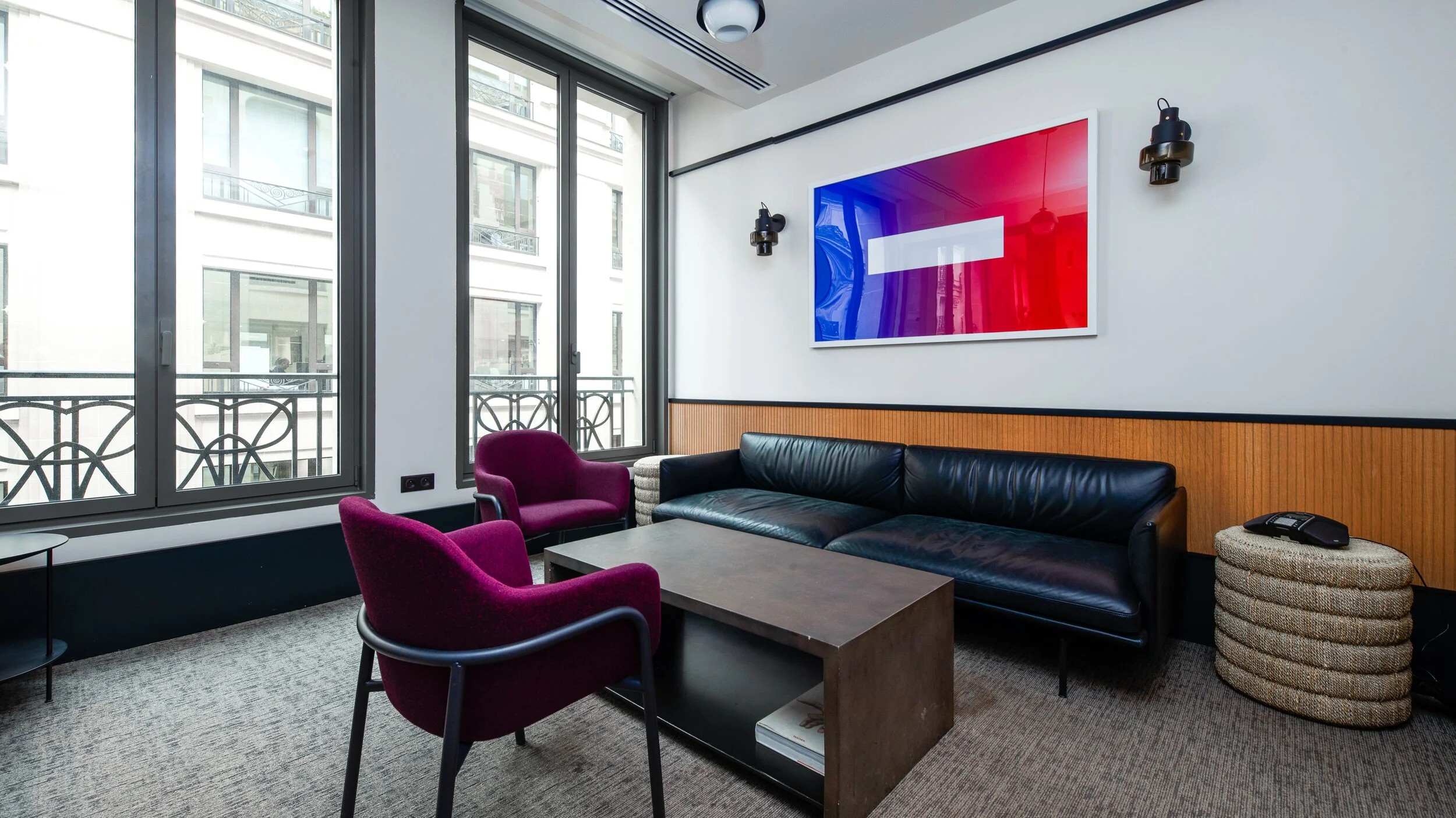 Modern office lounge area with black leather sofa, two purple armchairs, a wooden coffee table, and a large window showing apartment buildings outside. There is a framed red, blue, and white abstract art piece on the wall, with black wall-mounted lig
