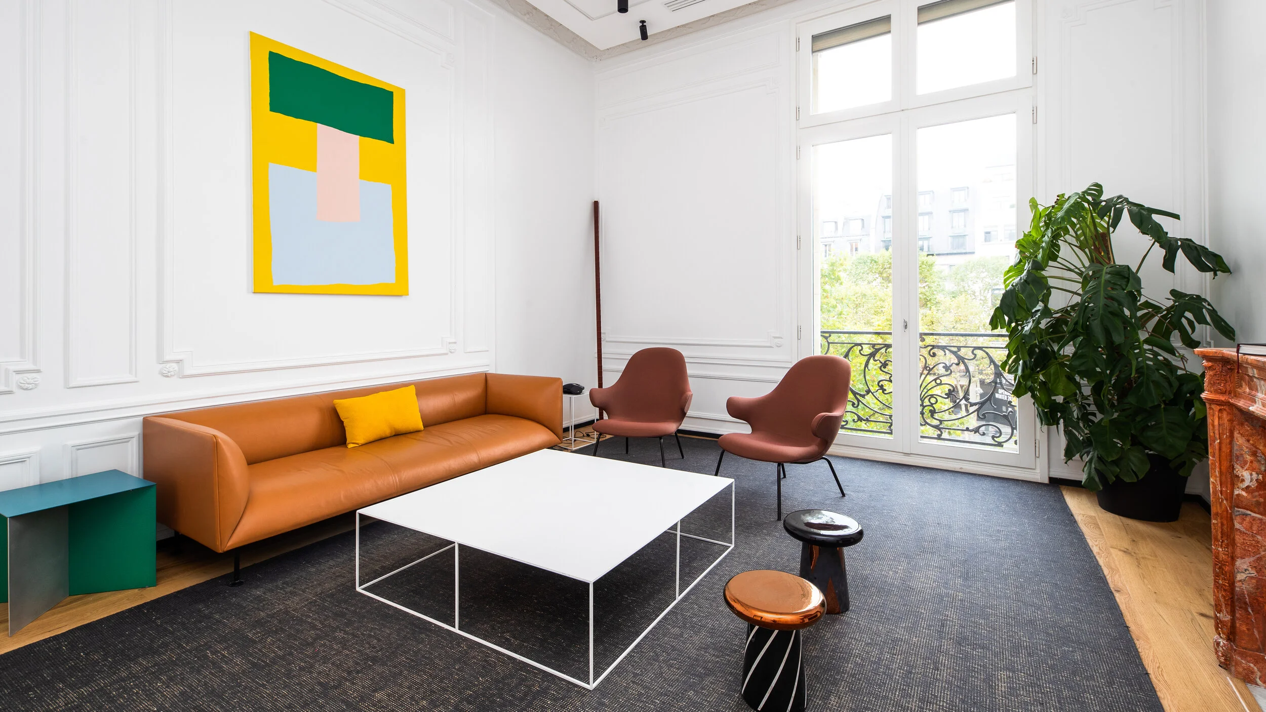 Modern living room with white walls, large windows, a brown leather sofa with yellow cushion, two pink chairs, a white coffee table, small stools, a potted plant, and colorful abstract artwork on the wall.