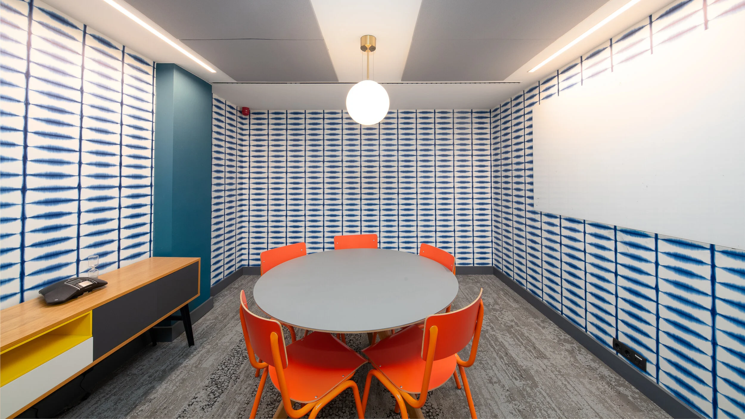 Conference room with a round table, orange chairs, a sideboard with a telephone and water glass, and wallpaper with a blue and white pattern. There is a pendant light hanging from the ceiling and electrical outlets on the wall.