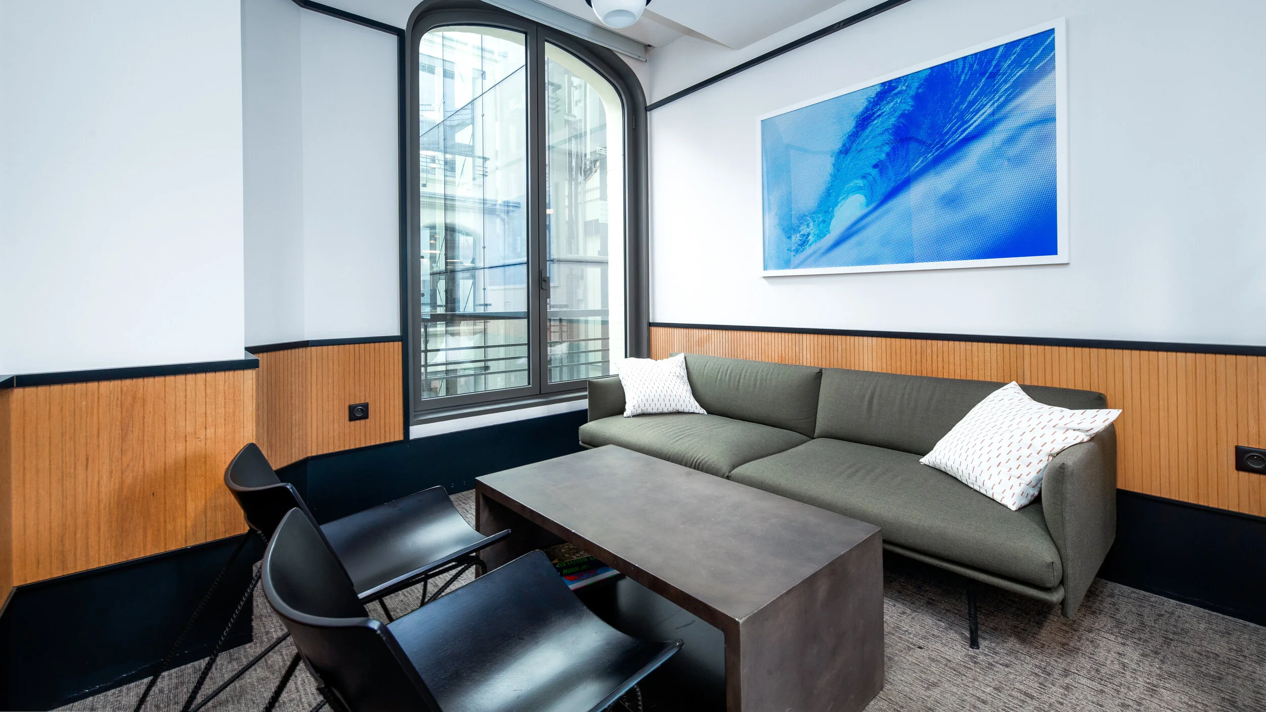 Modern office lounge with a gray sofa, two decorative pillows, black chairs, a concrete coffee table, large window with a view of a building, and a blue abstract artwork on the wall.