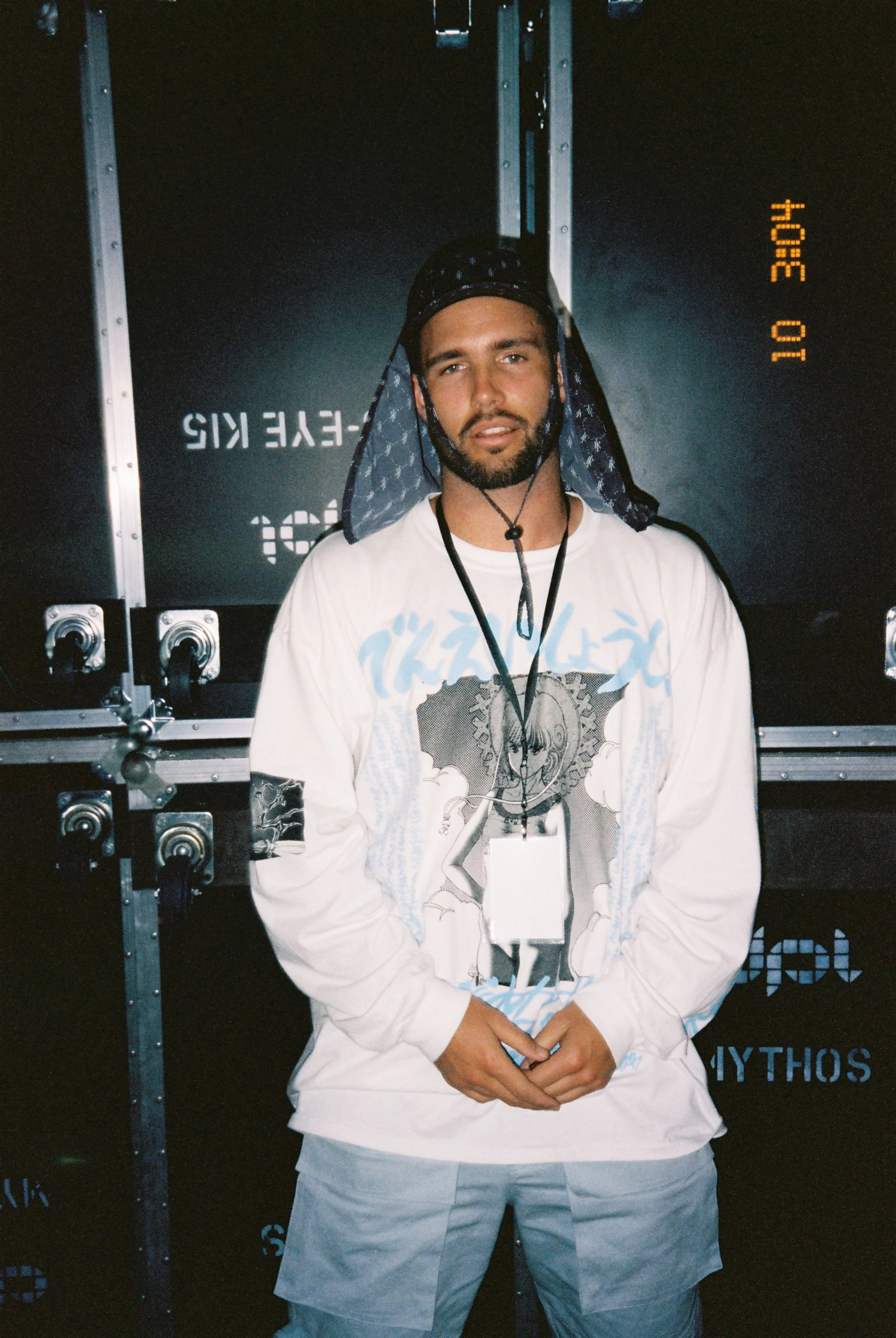 A man with a beard wearing a white long-sleeve shirt with graphic design, light gray shorts, a black lanyard with an ID badge, and a dark blue patterned head covering, standing in front of a black background with metal hardware and numeric digital da