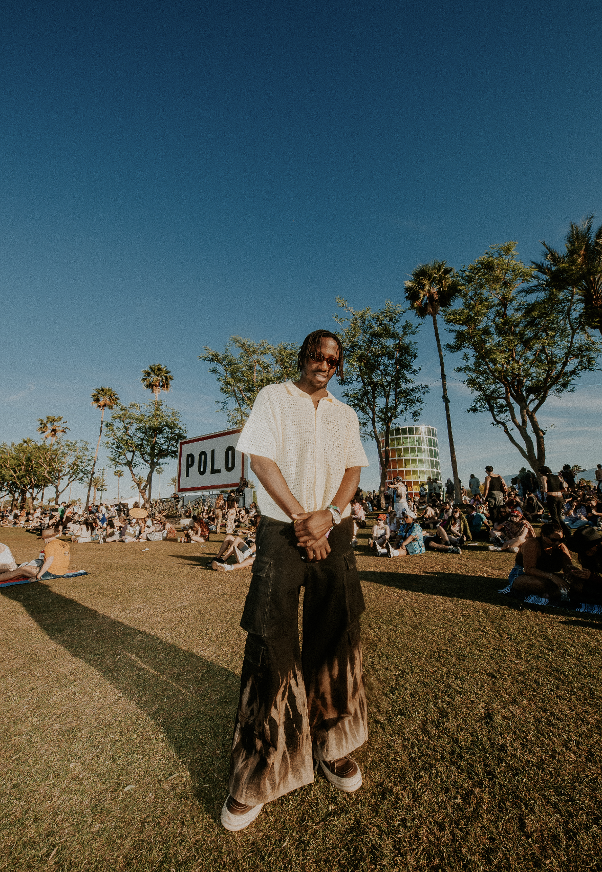 A man standing on a grassy field at an outdoor event with a crowd sitting around, tall palm trees, a large sign that says 'POLO', and a colorful building in the background under a clear blue sky.