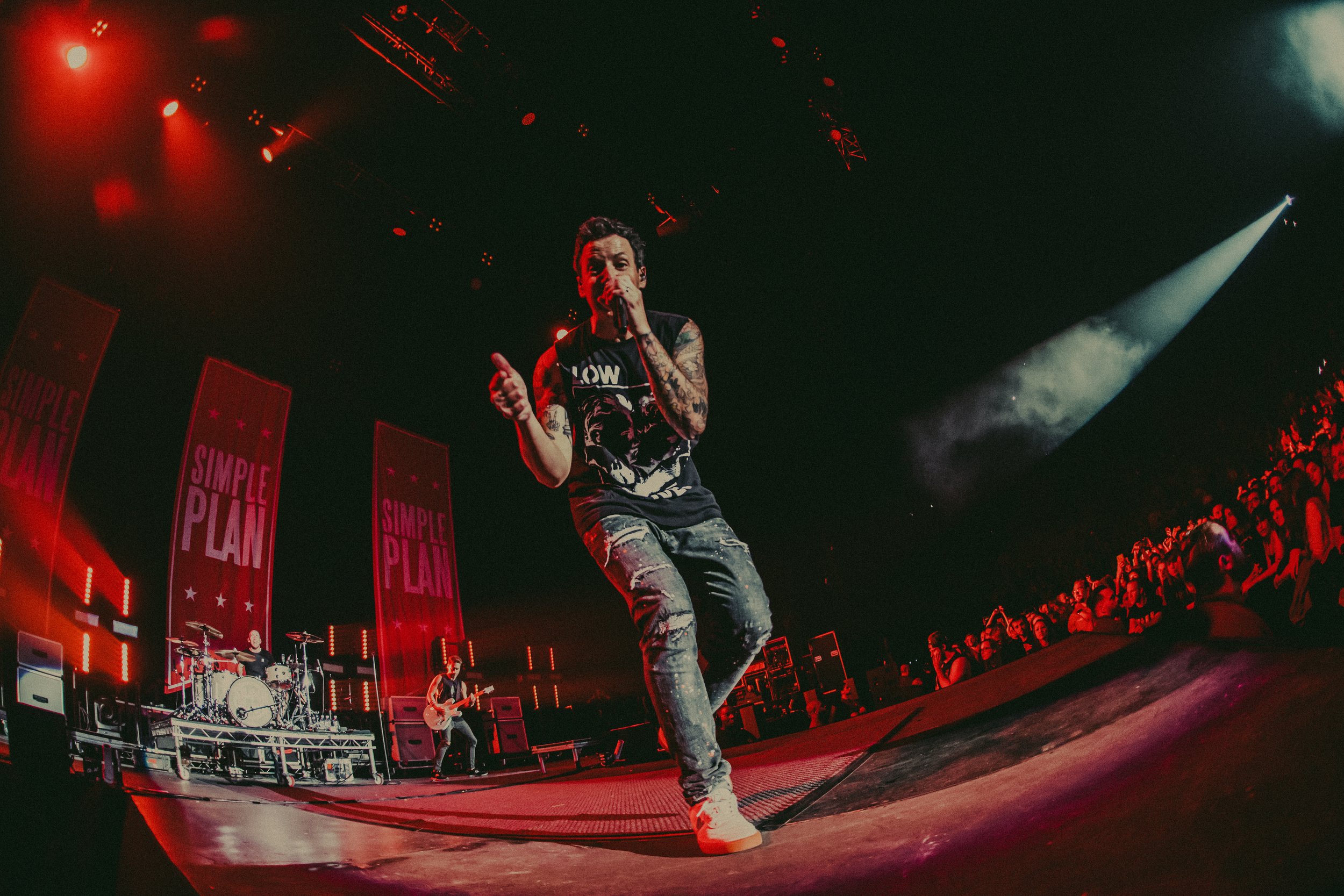 Male musician with tattoos singing into a microphone on stage during a concert. Behind him, a band with drum set and guitar, large vertical banners with 'SIMPLE PLAN' written on them, and an audience Watching the performance. Bright stage lights and 