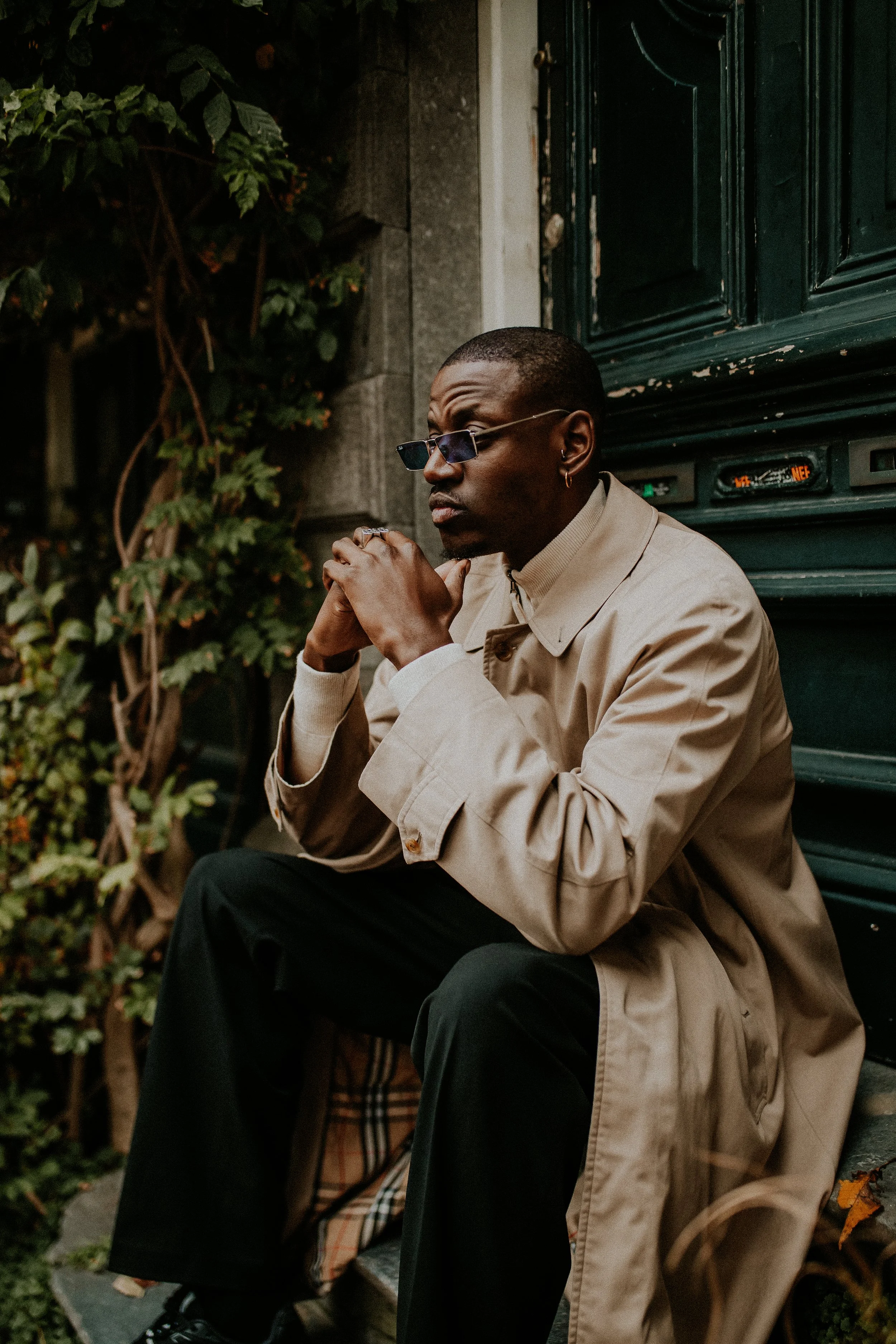 A man sitting on a step outside, wearing sunglasses, a beige trench coat, and black pants, with his hands clasped under his chin, looking off to the side, surrounded by greenery and a dark green door