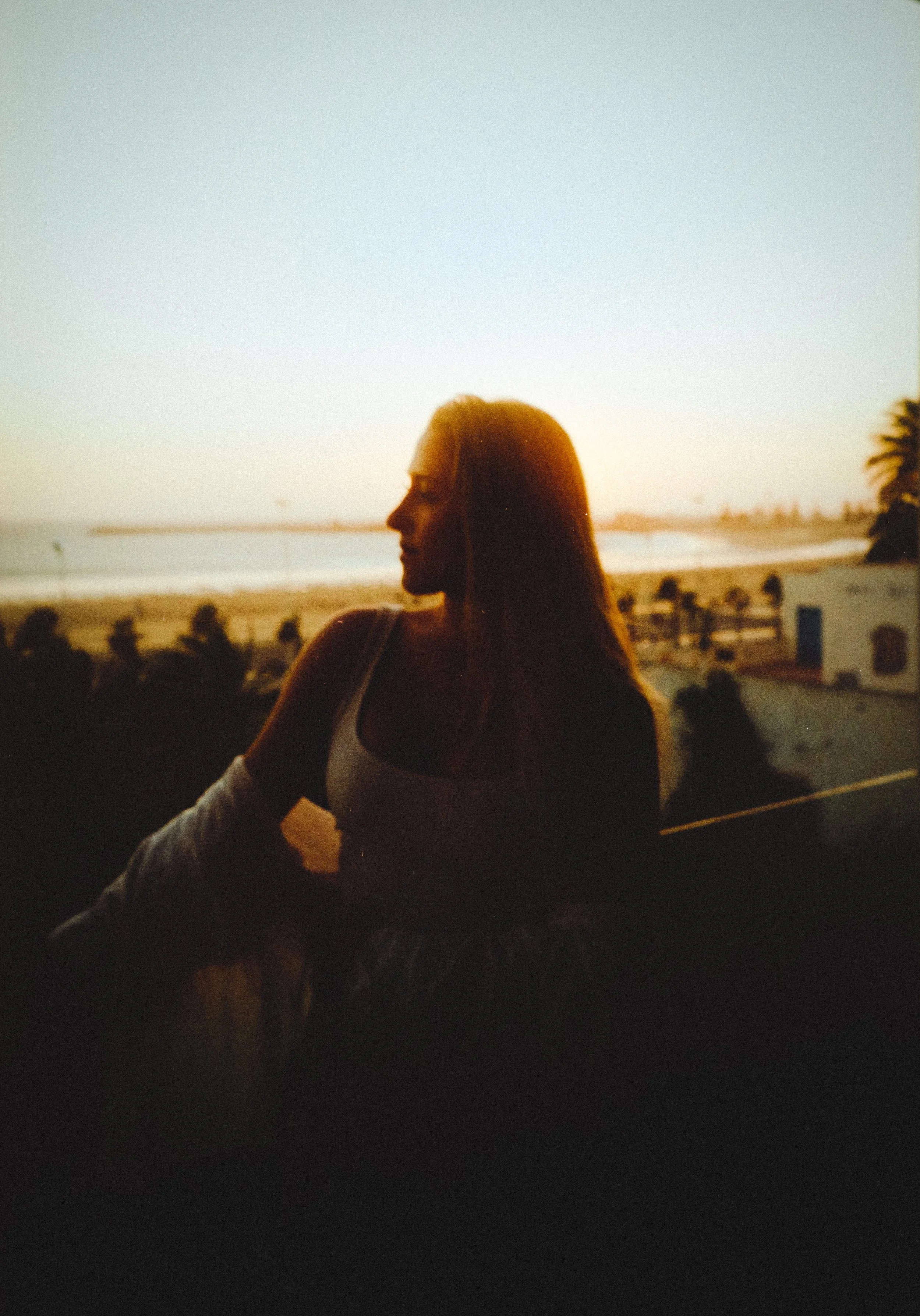 Silhouette of a woman with long hair looking to the side during sunset by the beach, with trees and buildings in the background.