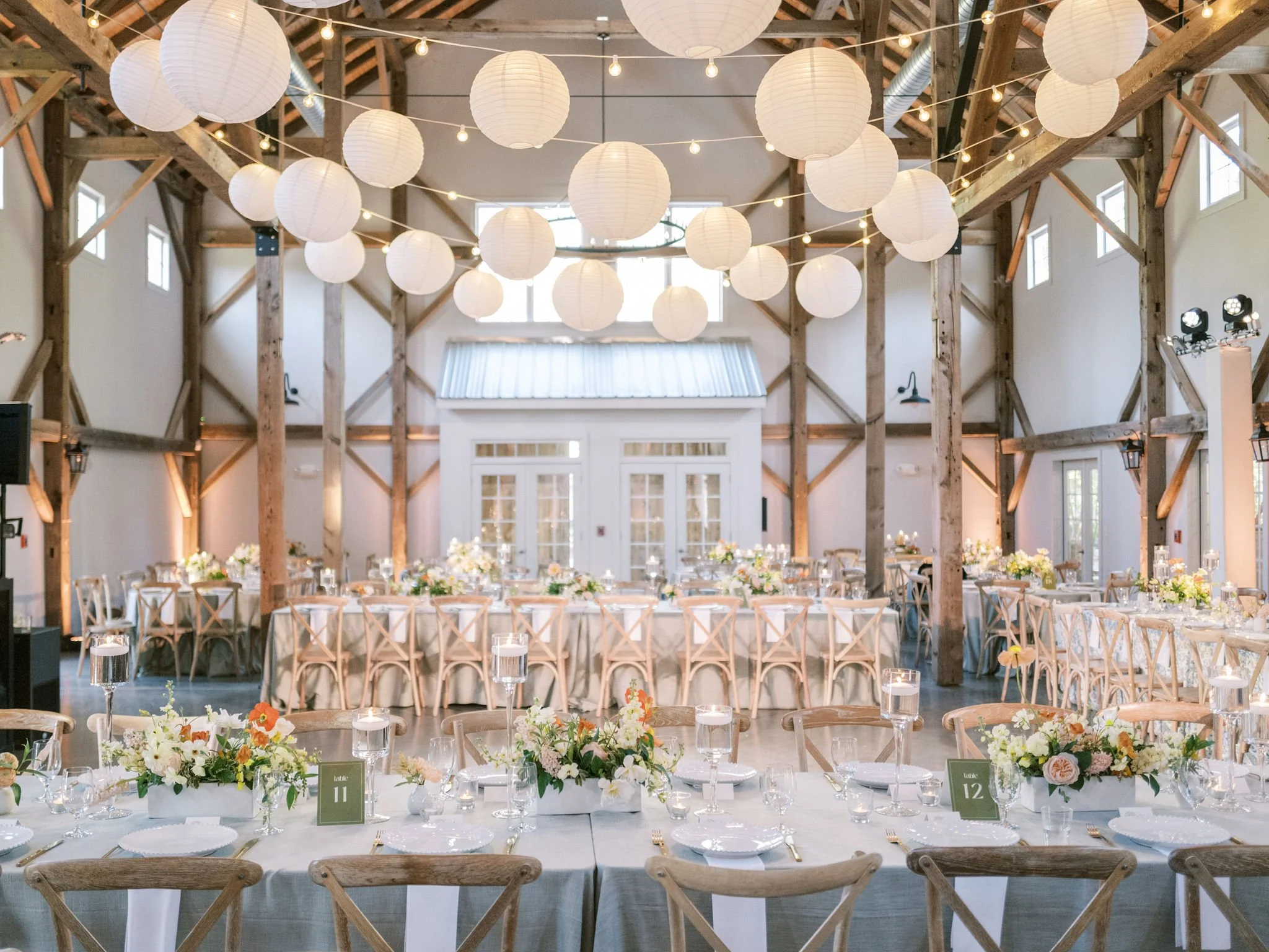 Wedding reception inside The Barn of Chapel Hill with wooden beams, string lights, and white paper lanterns above decorated tables