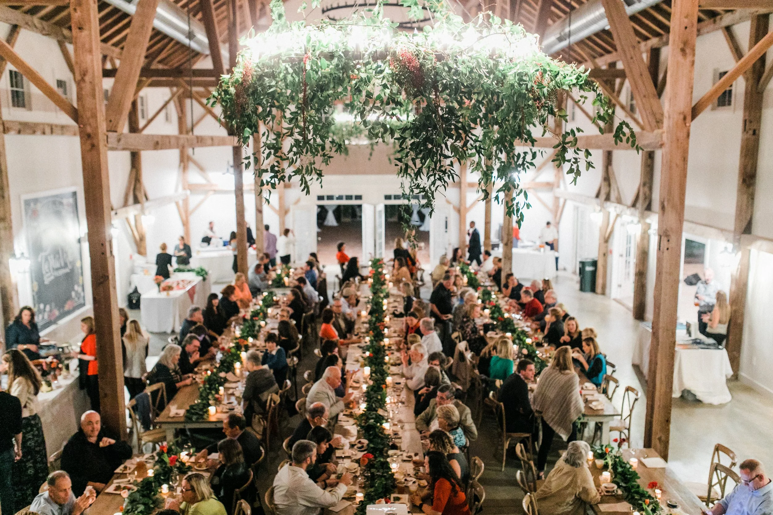 Guests gathered for a large dinner event inside The Barn of Chapel Hill beneath a hanging greenery installation