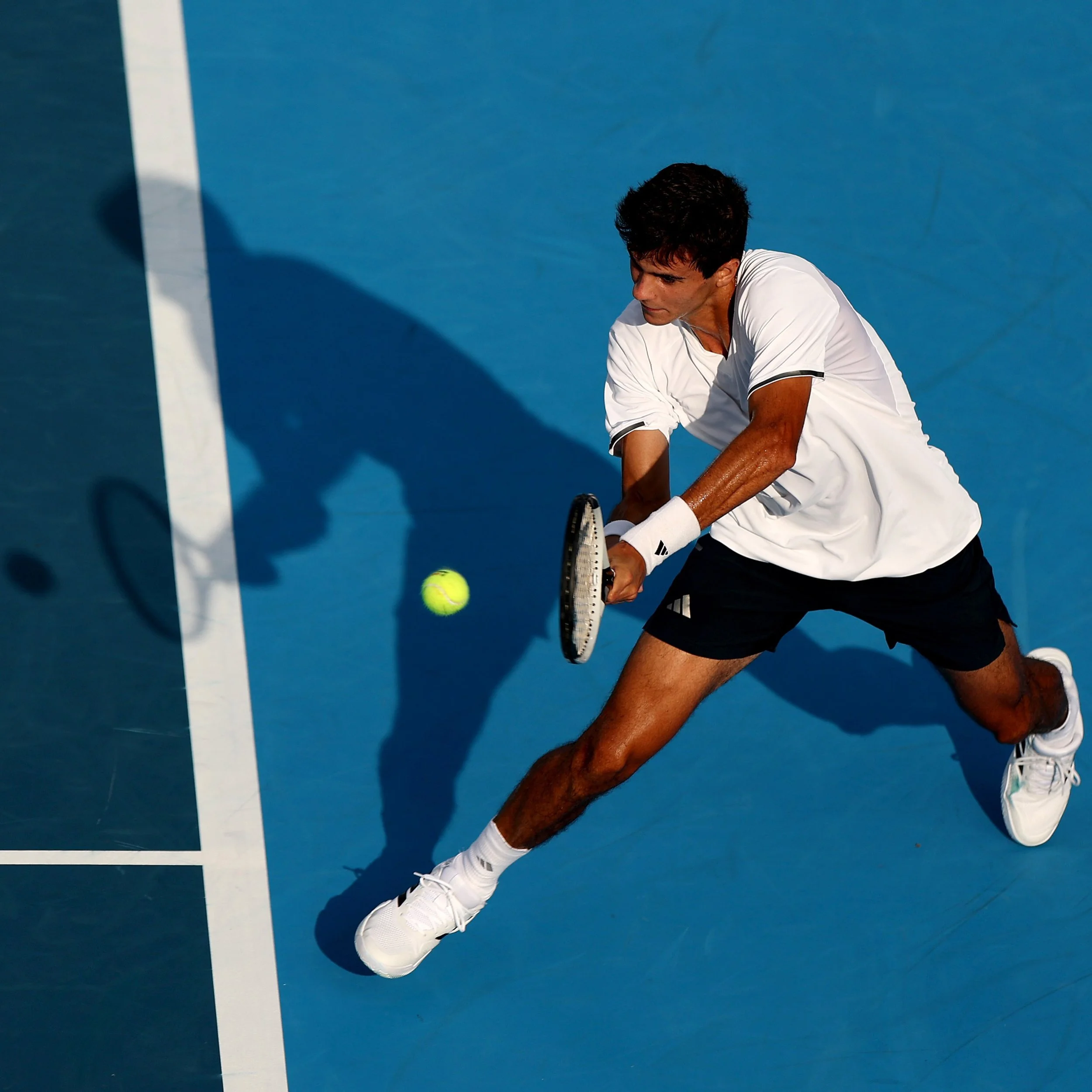 A male tennis player in white sportswear is preparing to hit an incoming tennis ball on a blue court.