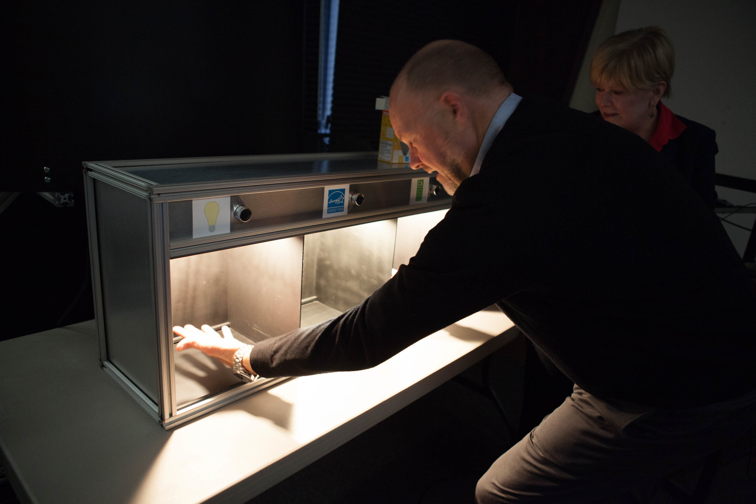 A man with short hair wearing a black sweater over a blue dress shirt comparing an incandescent light to an Energy Star-rated light in a light box..jpg