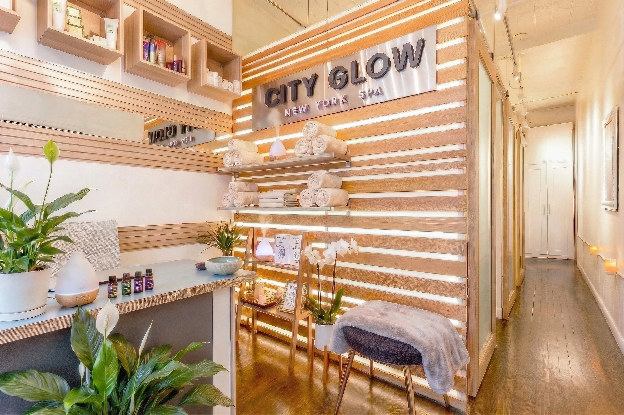 Interior of a spa with wooden decor, shelves with towels and skincare products, a sign reading 'City Glow New York Spa,' plants, and warm lighting