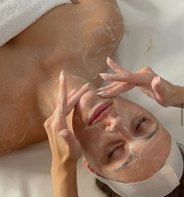 A woman lying down with her eyes closed, receiving a facial treatment while a skincare specialist applies a facial mask or cleanser to her face.
