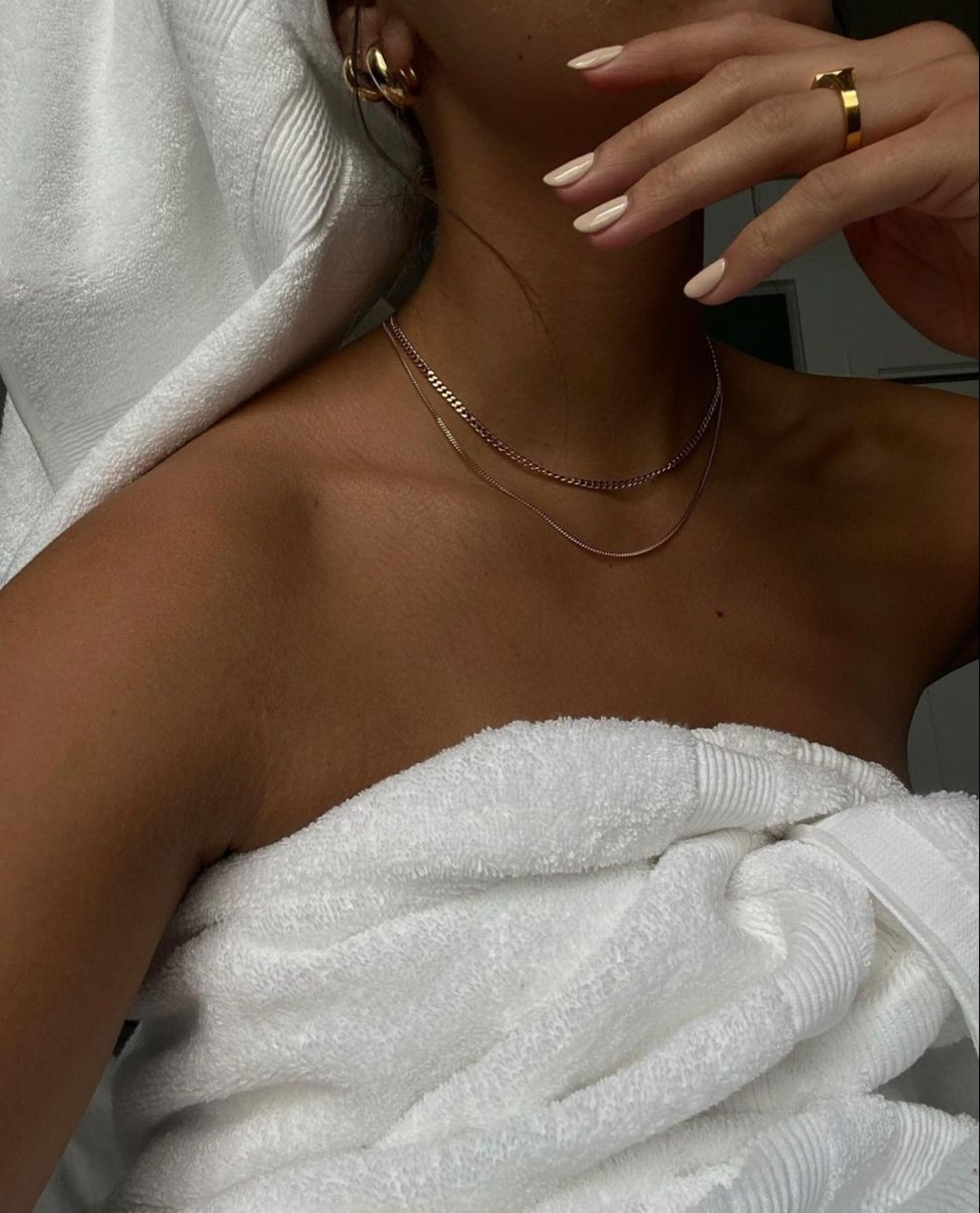 A woman lying in bed wrapped in a white towel, wearing jewelry including layered necklaces, earrings, and rings, with her hand near her face and her face partially visible.