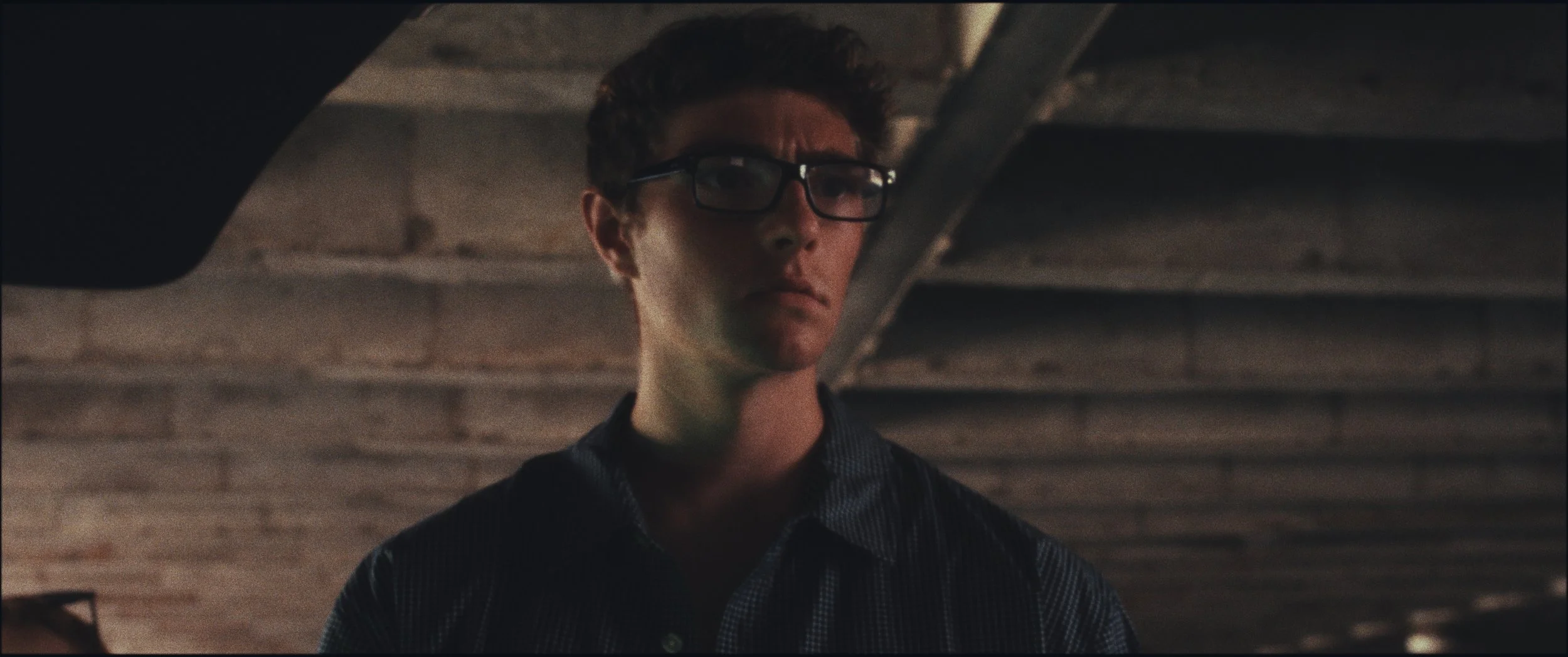 Low-angle shot of a man in glasses in a parking garage, still from Five Finger Stakeout (2026), shot in Omaha, Nebraska by cinematographer Cole Mowery