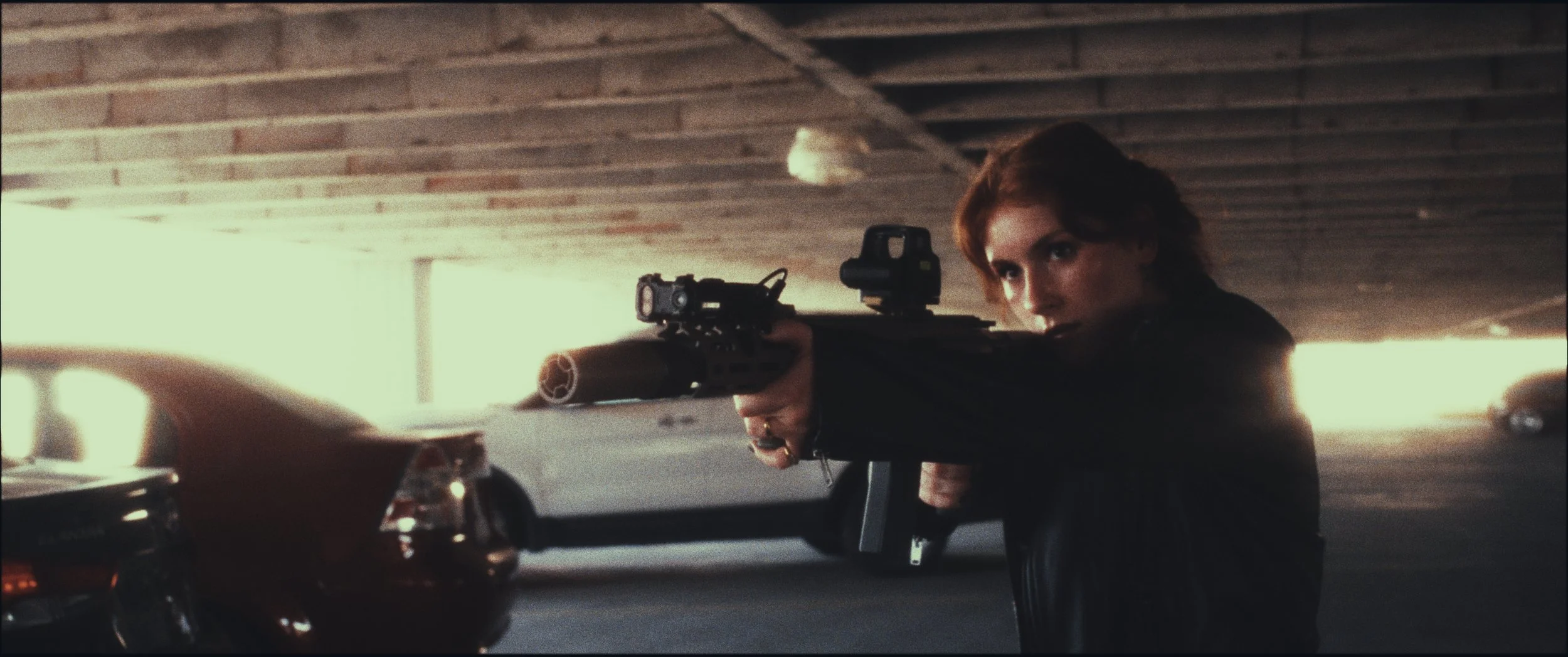 Woman aiming a rifle in a parking garage, still from Five Finger Stakeout (2026), shot in Omaha, Nebraska by cinematographer Cole Mowery
