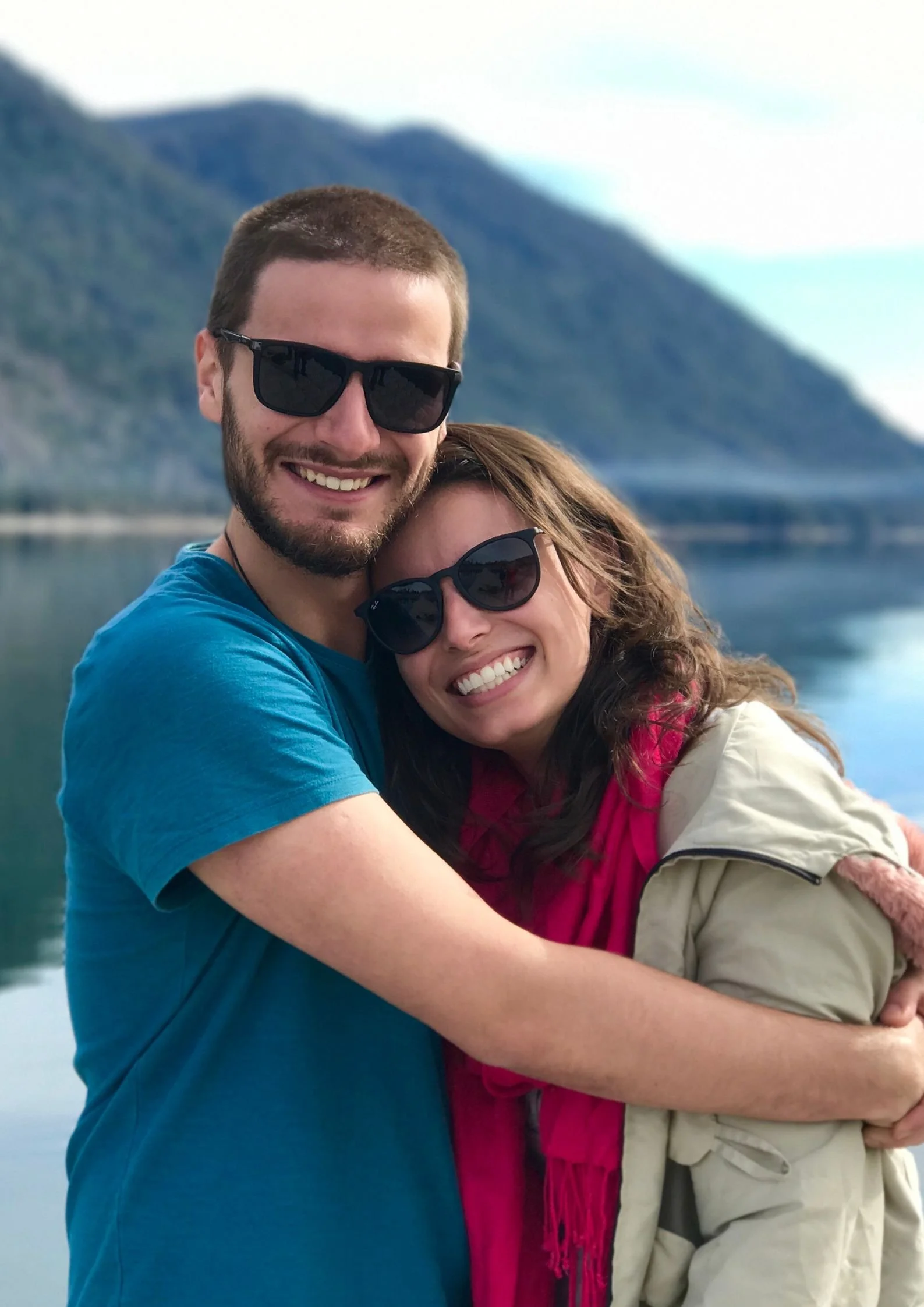 Una coppia felice si abbraccia davanti a un lago con montagne sullo sfondo, entrambi indossano occhiali da sole e sorridono.