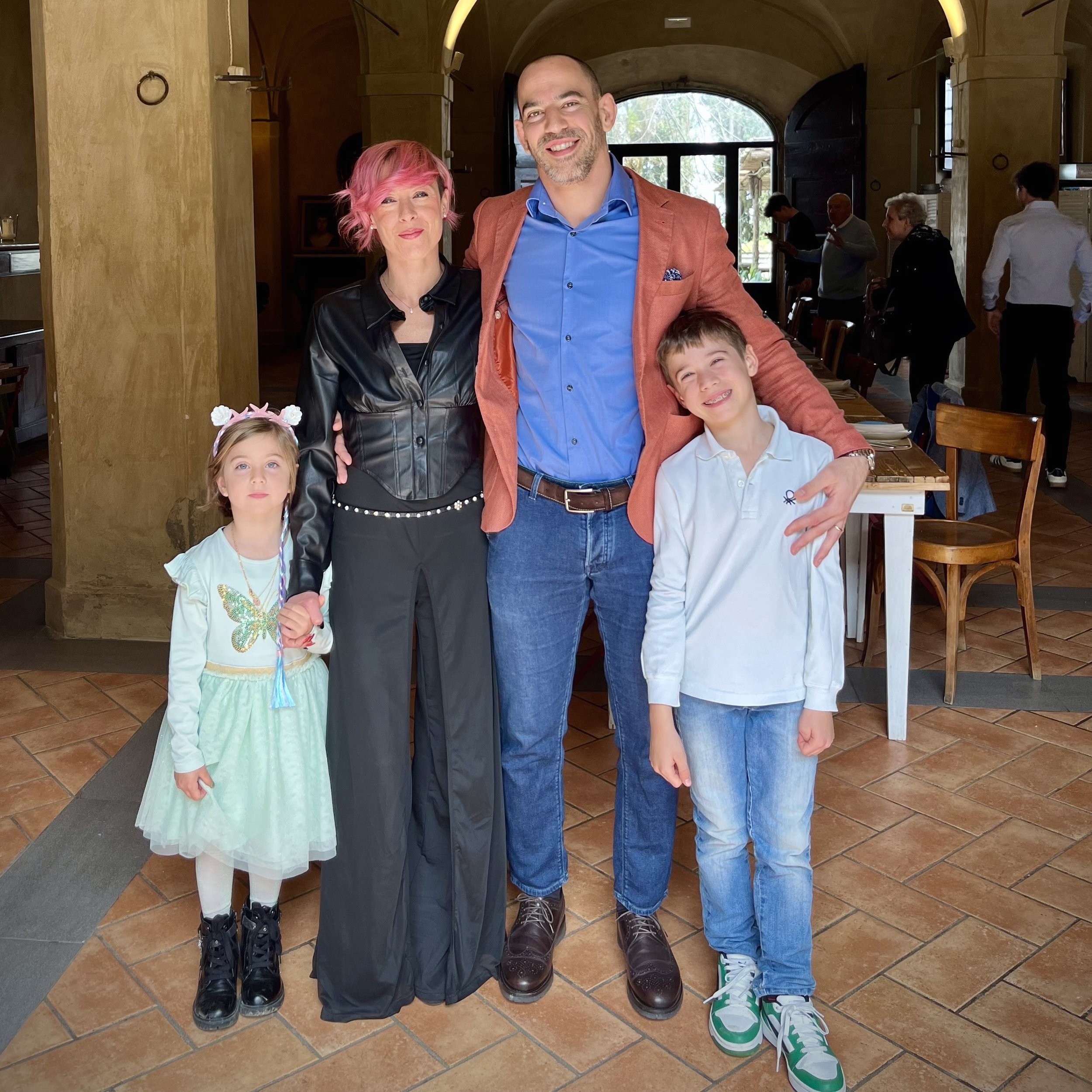 Una famiglia di cinque persone davanti in un ristorante, con tre adulti e due bambini, tutti sorridenti e vestiti casual eleganti.
