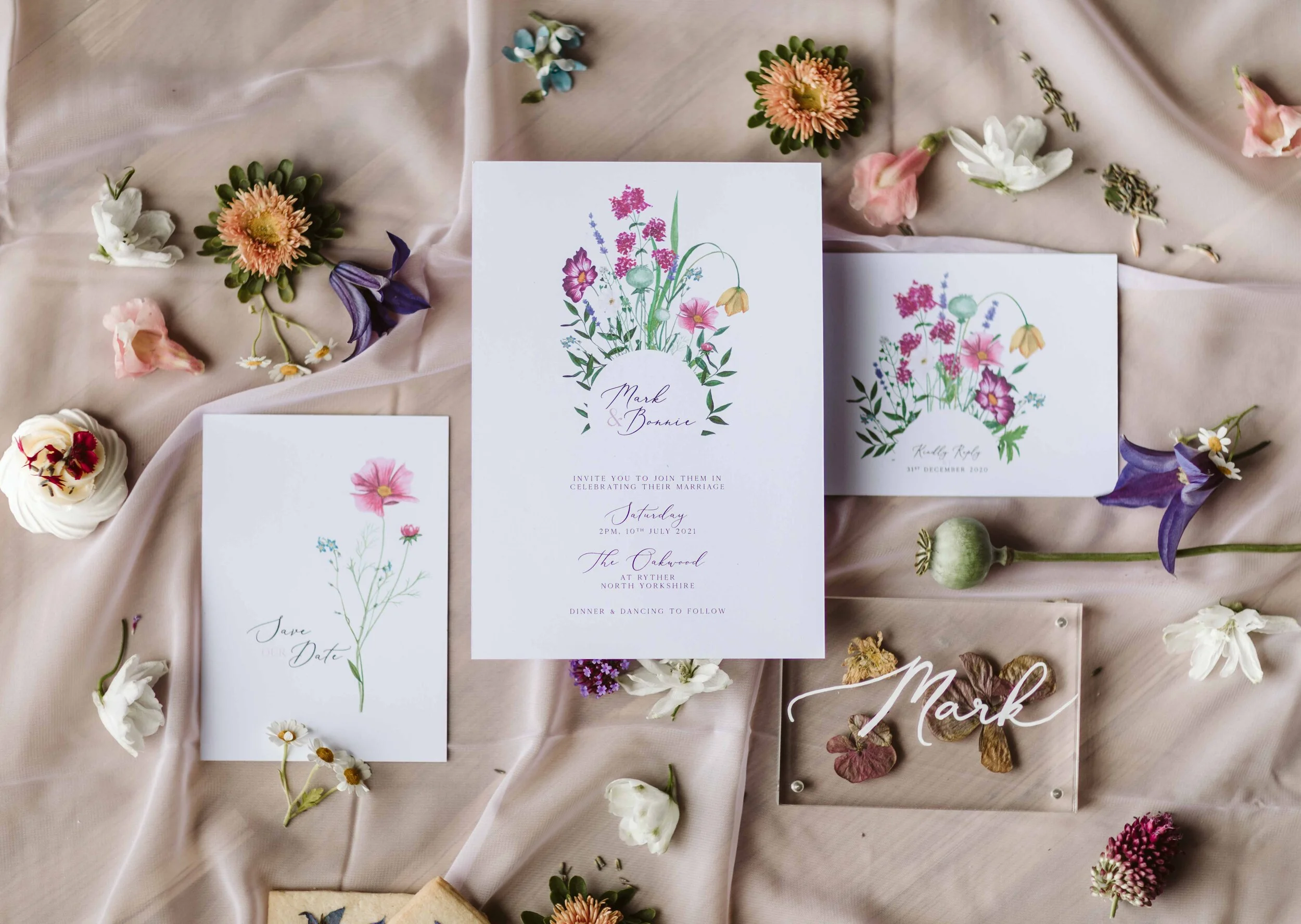Wedding invitation cards with floral designs surrounded by various flowers and petals on a soft cloth background.