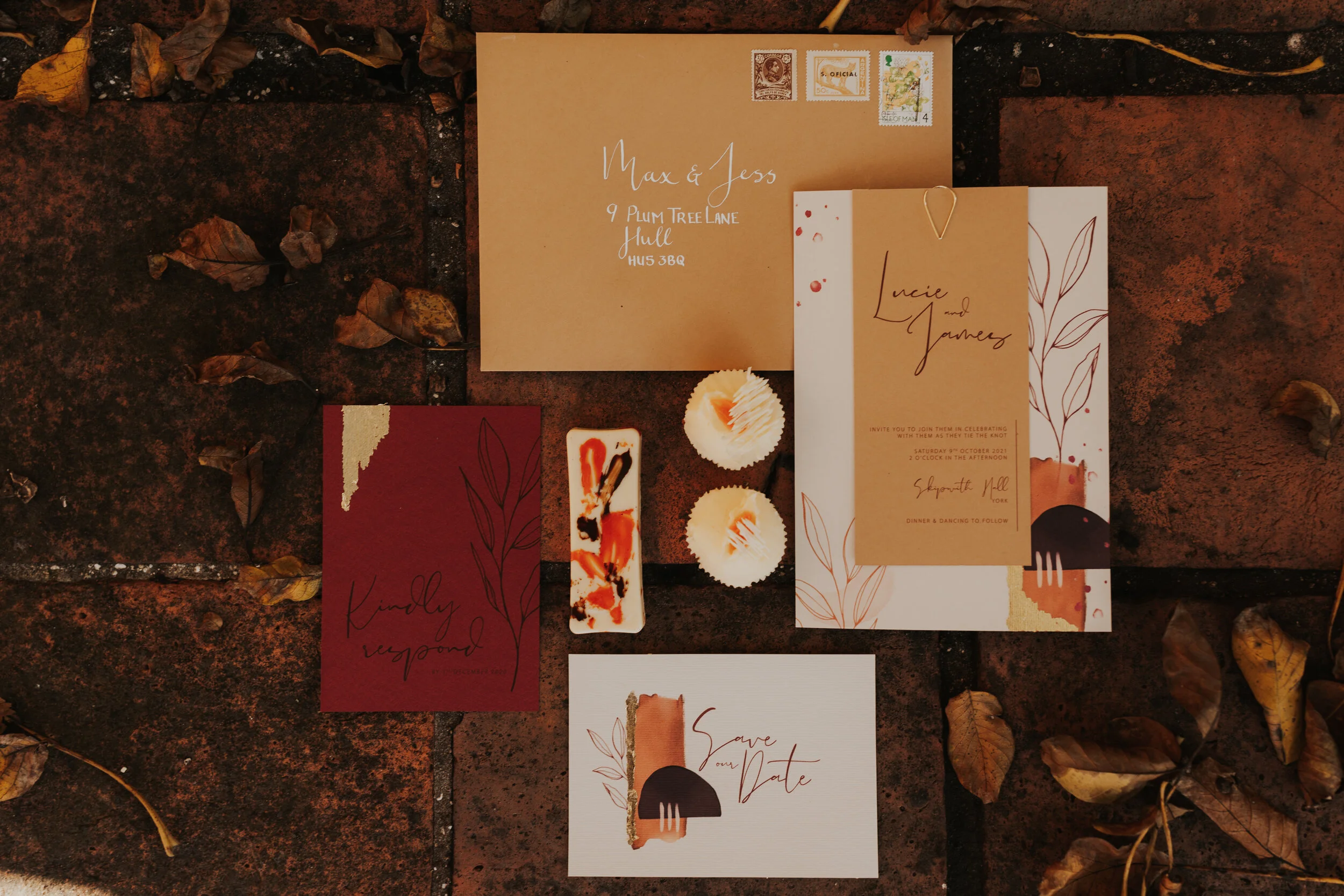 Set of wedding stationery including invitations and RSVP cards with earthy tones and minimalist leaf designs, placed on a brick surface with dried leaves around.
