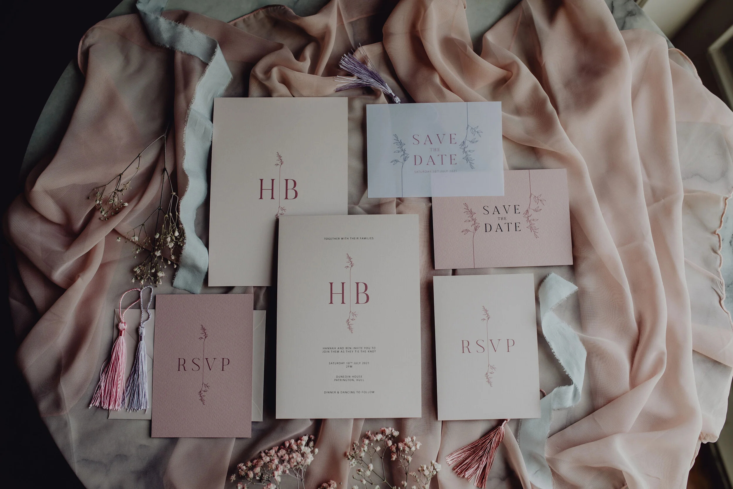 Set of wedding stationery including save the date cards, RSVP cards, and an invitation, arranged on soft pink and beige fabrics with some small flowers and tassels as decorative items.