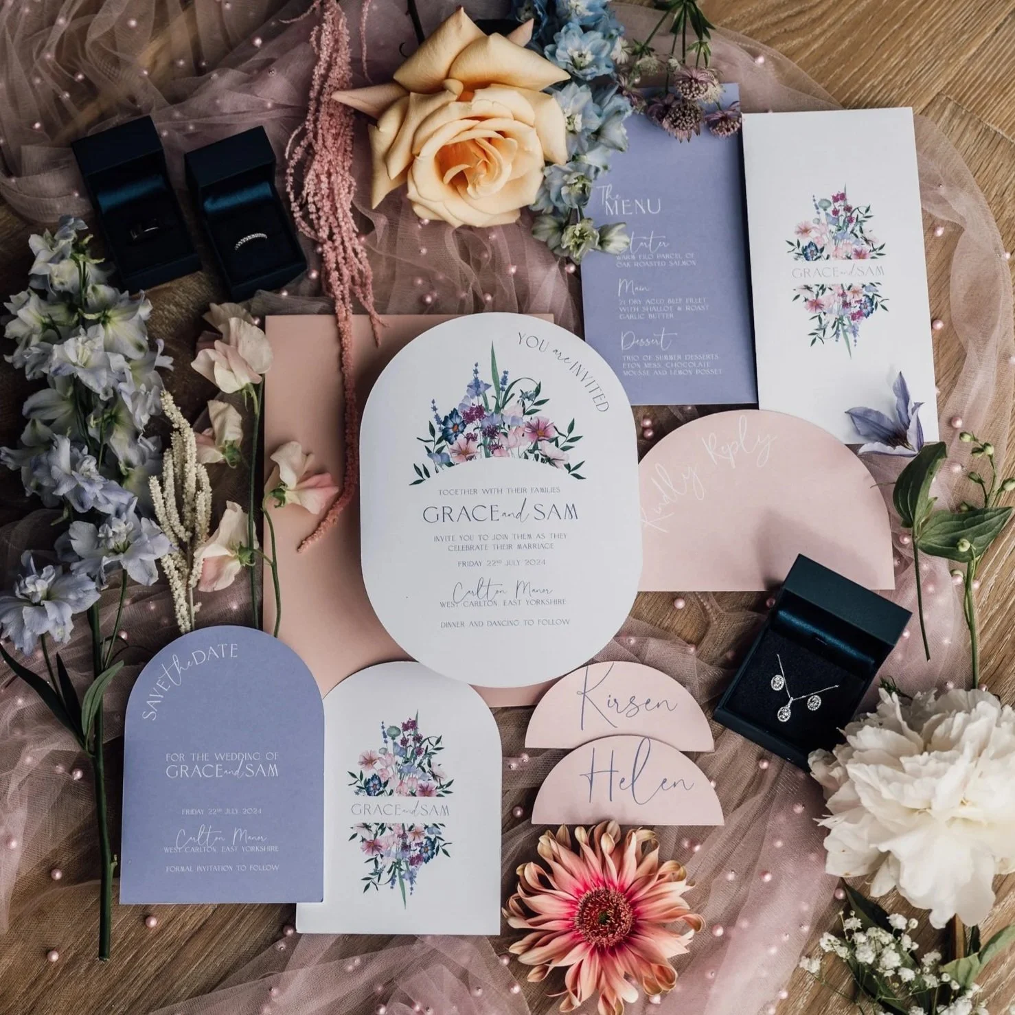 Wedding invitations and save the date cards decorated with pink, purple, and white flowers, along with jewelry boxes containing rings and a necklace. Various flowers and pink beads are arranged around the stationery on a wooden surface.