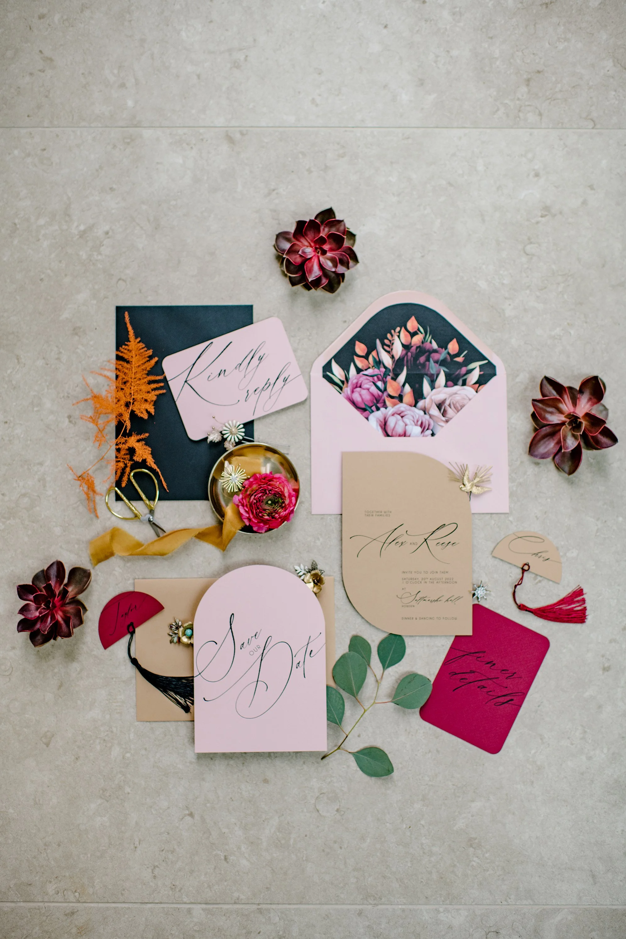 A flat lay of elegant wedding invitations, save-the-date cards, and RSVP cards with floral and botanical accents, surrounded by small succulent plants and decorative elements. The invitations feature cursive calligraphy and pastel colors.