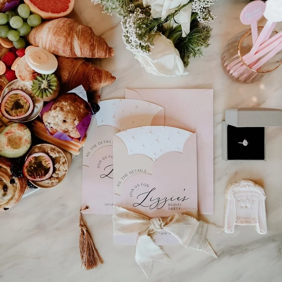 A bridal shower invitation with pink and white accents. Surrounding items include assorted pastries, fruit, a bouquet of white flowers, a ring in a box, pink cake toppers, and decorative baby onesie and bow accessories.
