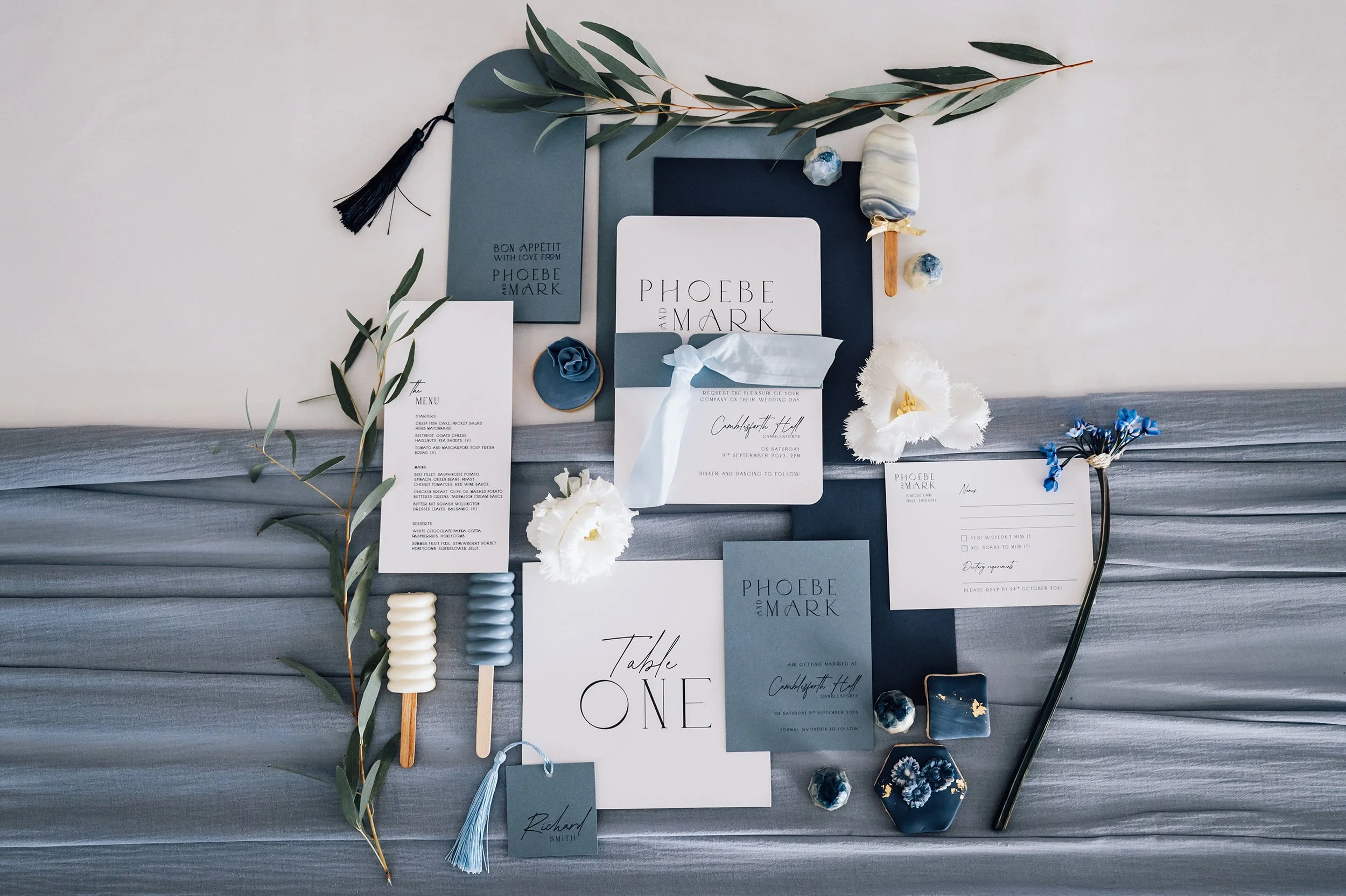 Wedding invitation suite with stationery, flowers, and decorative items in white, blue, and gray tones arranged on a gray fabric surface.