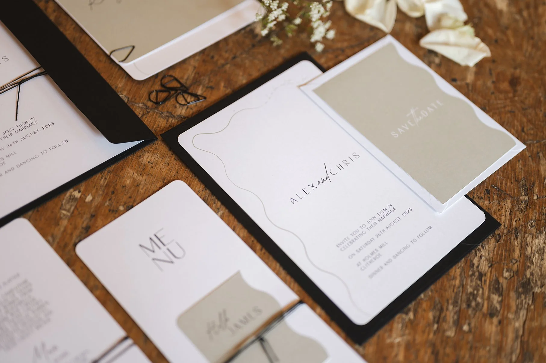 Wedding invitations and save-the-date cards on a wooden table with paper clips, a white box, and flower petals.