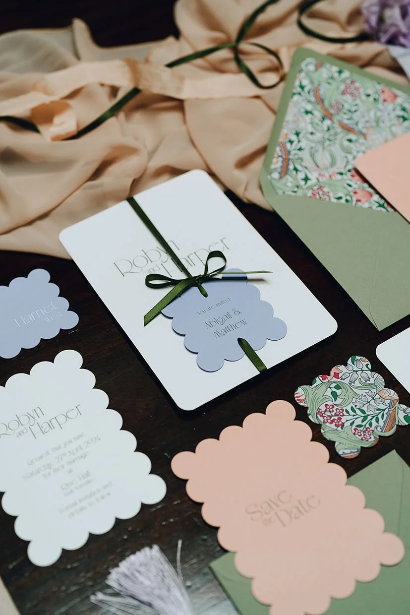 Photo of a wedding invitation suite with matching RSVP card, save the date card, and decorative paper embellishments on a dark wooden surface, surrounded by a beige fabric with green ribbon detail.